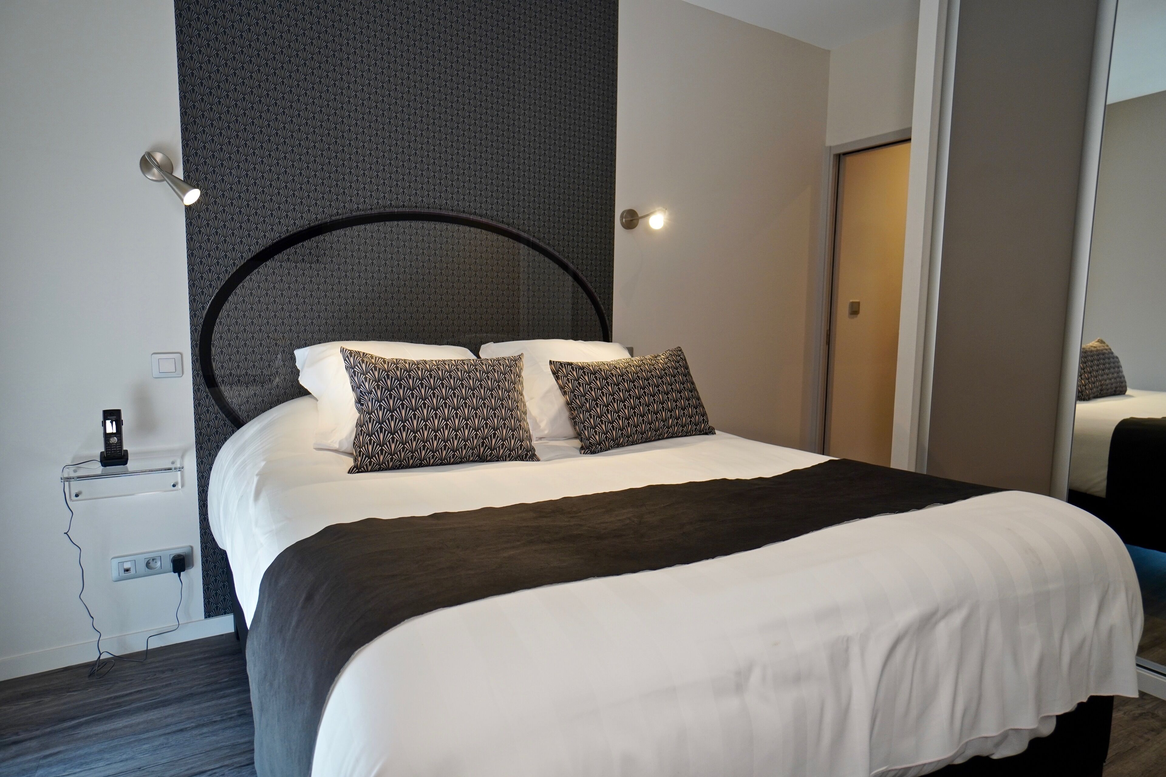 A neatly made bed with patterned pillows, a bedside table with a lamp, and a power outlet.