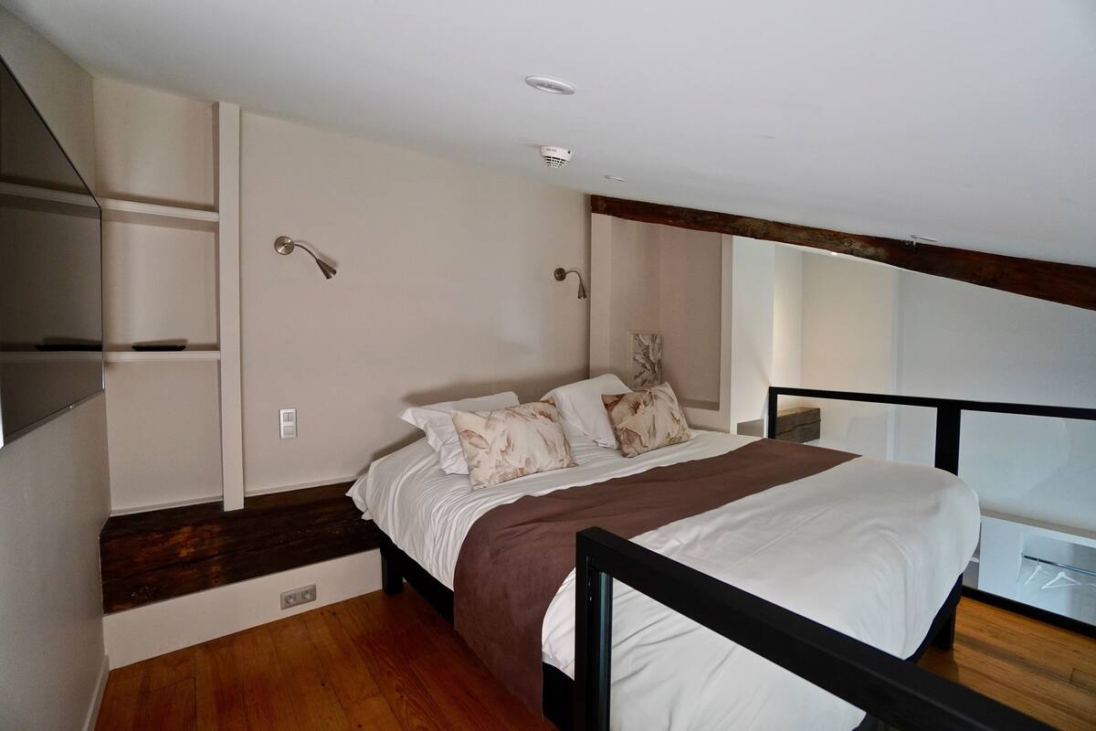 A bedroom with a bed, wooden flooring, a brown accent wall, and a window with a view.