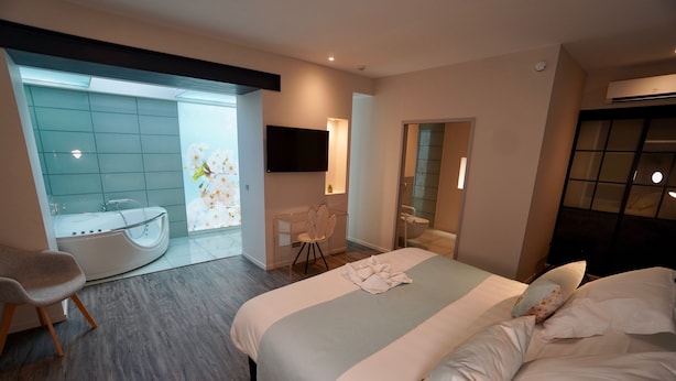 A modern hotel room with a large bed, a sitting area with a chair, a bathtub, and a bathroom with a mirror and sink.
