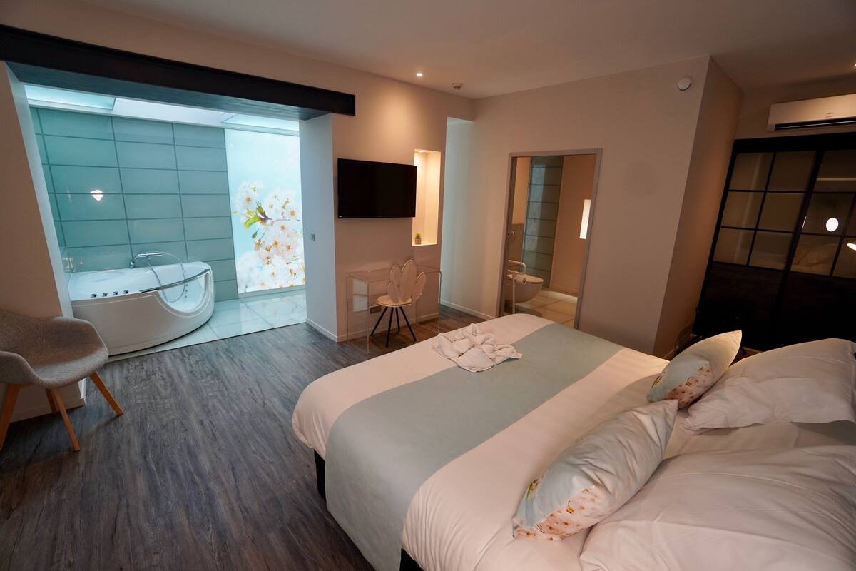 A modern hotel room with a large bed, a sitting area with a chair, a bathtub, and a bathroom with a mirror and sink.