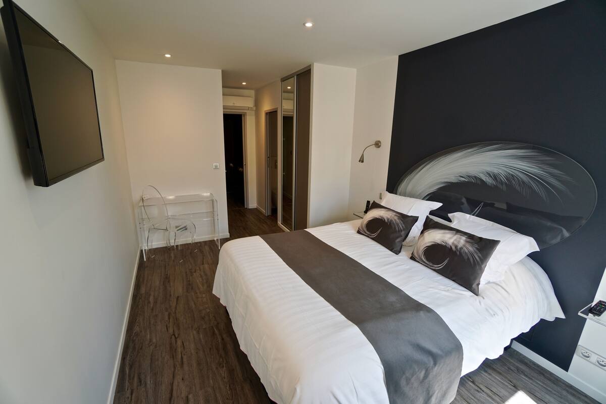 A modern hotel room with a large bed, a flat-screen TV, a bedside table, and a mirror.