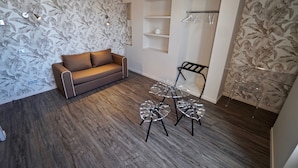 A room with a brown sofa, a small table with glass legs, and a wardrobe with hanging clothes.