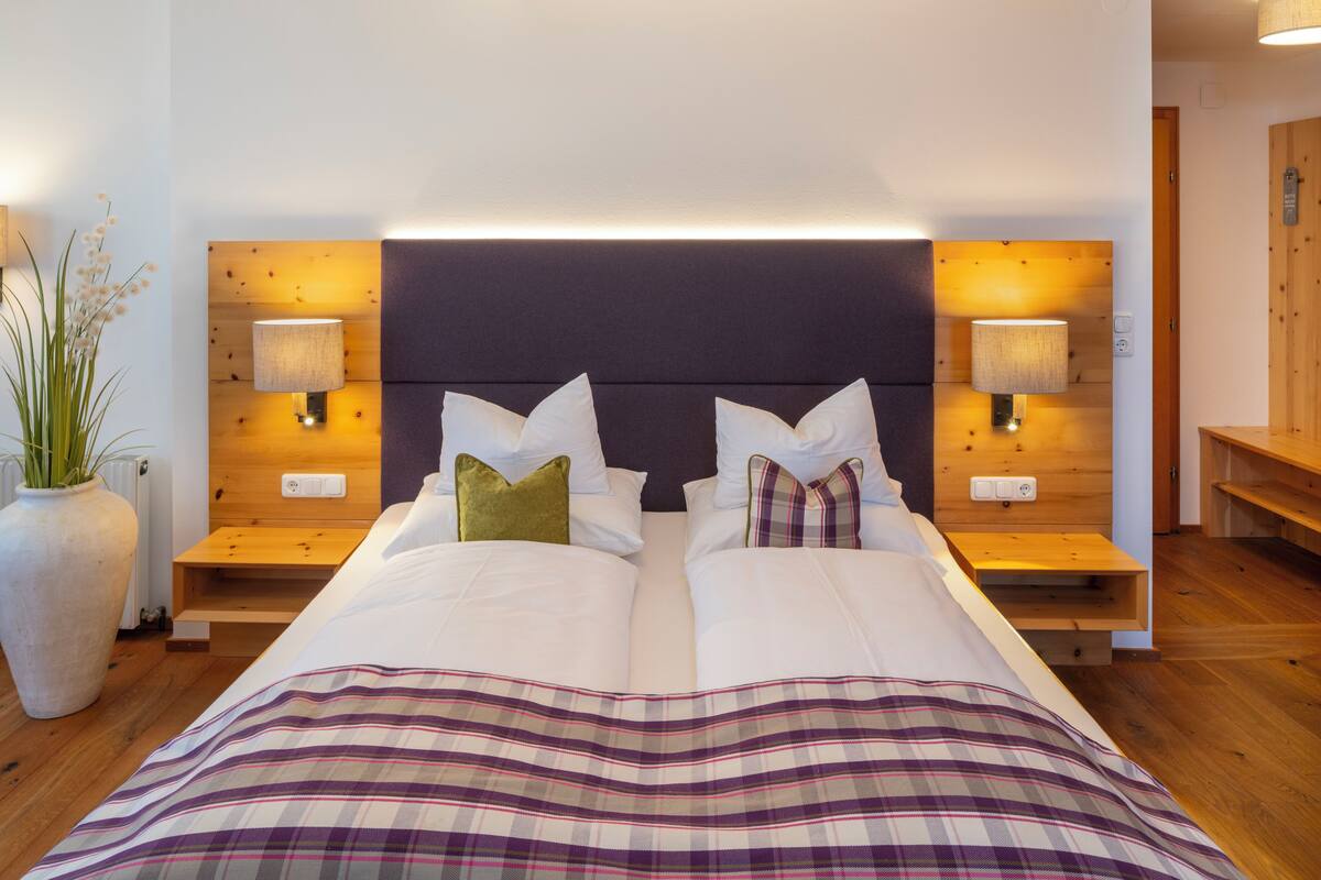 A double bed with a plaid bedspread, two bedside tables with lamps, a large potted plant, and a wooden headboard.