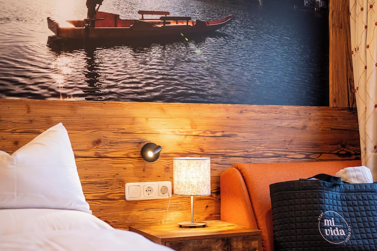 A hotel room with a bed, a nightstand with a lamp, a suitcase, and a view of a person rowing a boat on a lake.