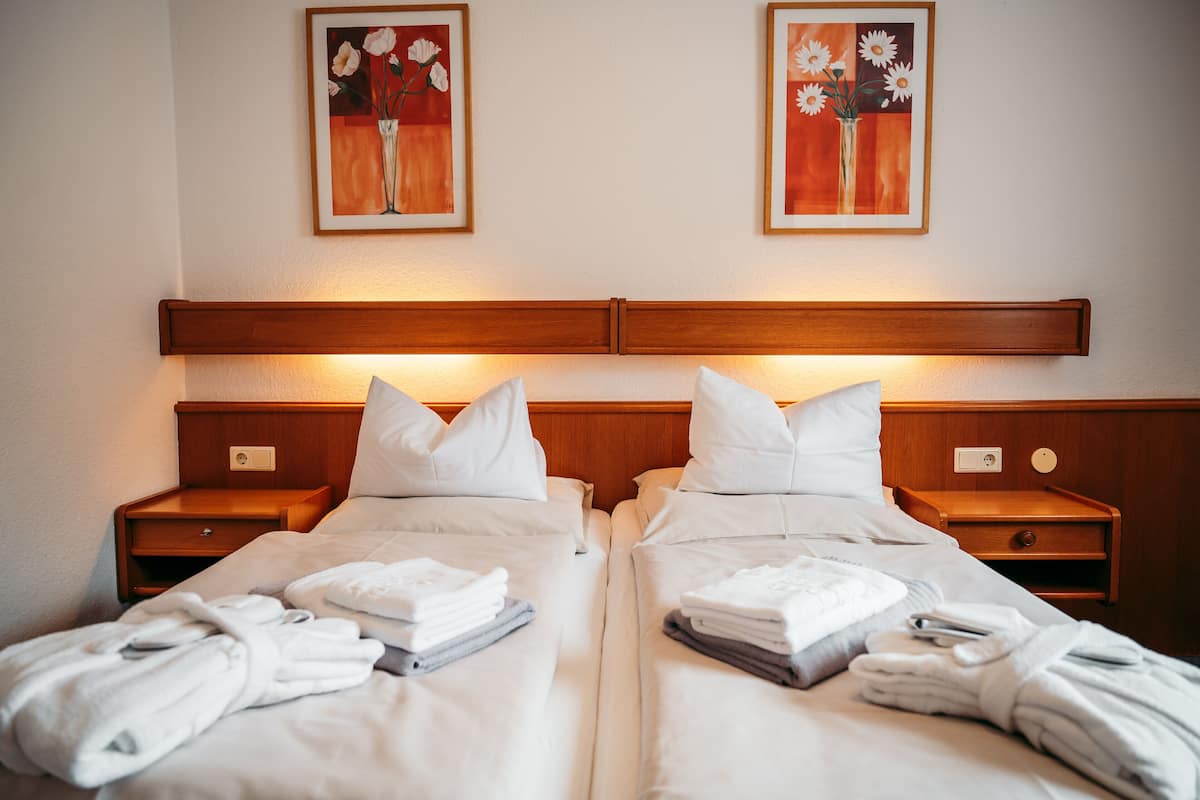 A hotel room with two beds, each with white linens and pillows, and bedside tables with lamps. There are framed floral paintings on the wall above the beds.