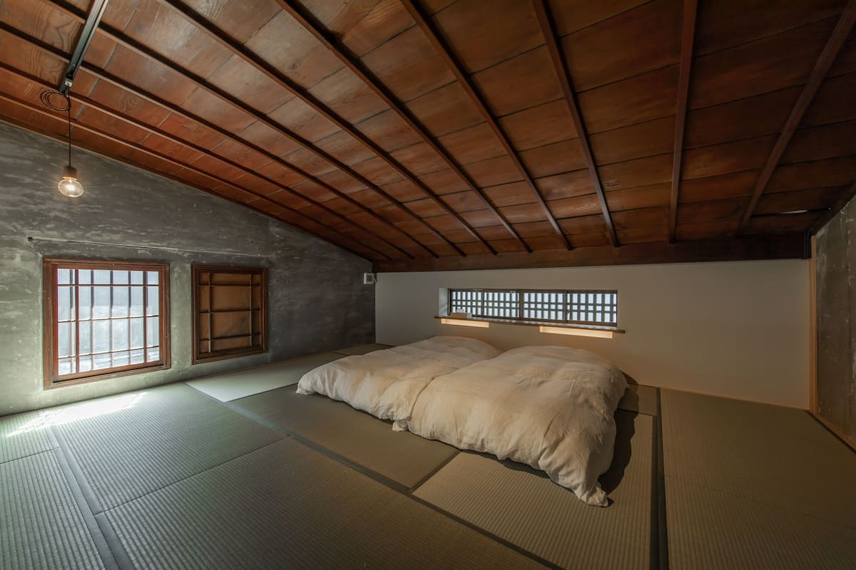 Ein traditioneller japanischer Raum mit Tatami-Böden, niedriger Decke mit Holzbalken und einem Bett mit weißer Bettwäsche.