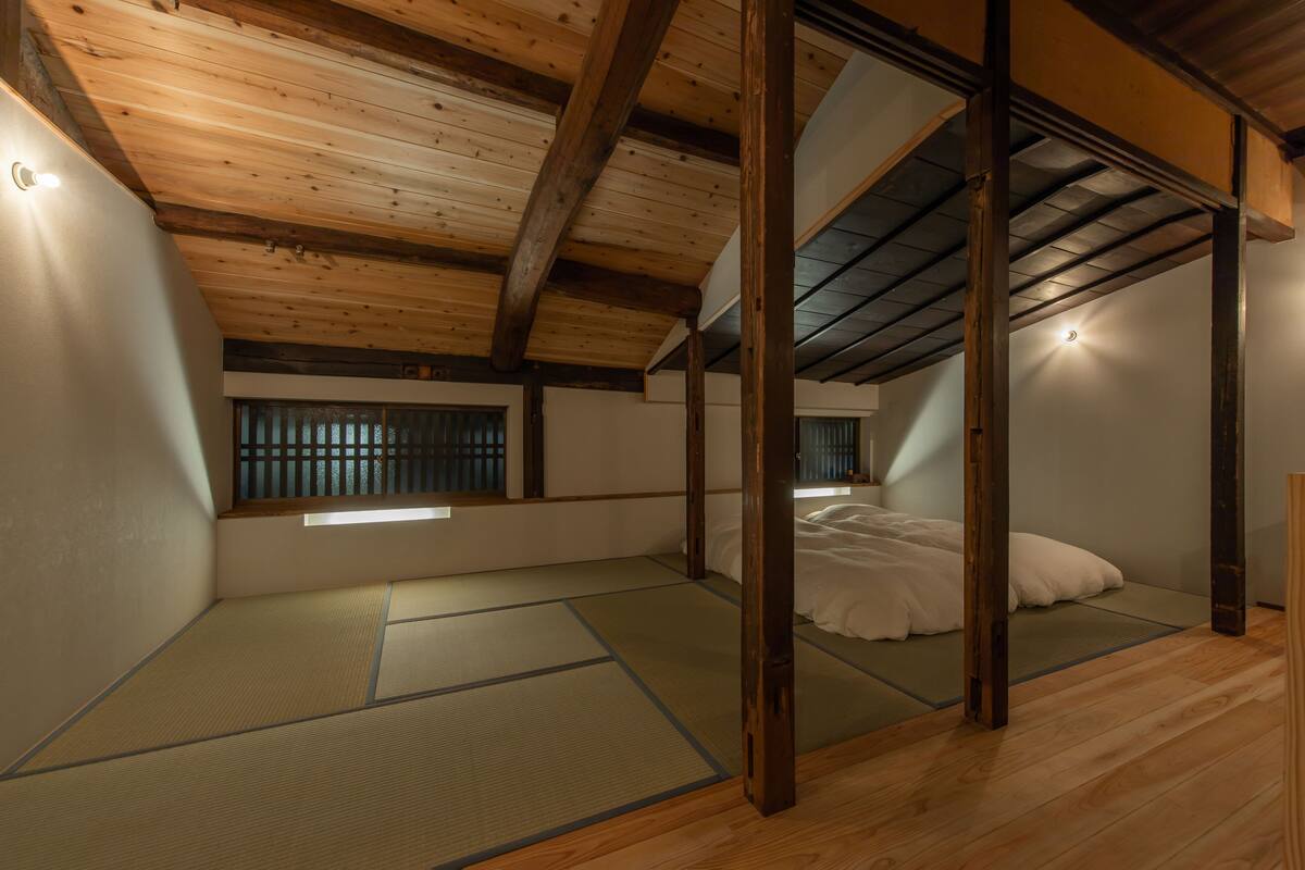 Ein traditioneller japanischer Raum mit Tatami-Boden, niedriger Decke und sichtbaren Balken sowie einem Bett mit weißer Bettwäsche.