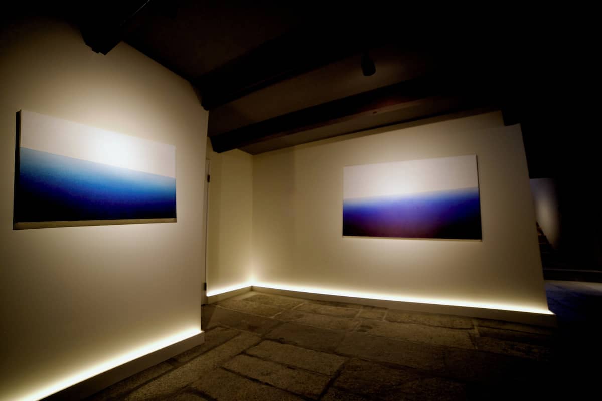Eine Kunstgalerie mit zwei beleuchteten Gemälden an weißen Wänden.