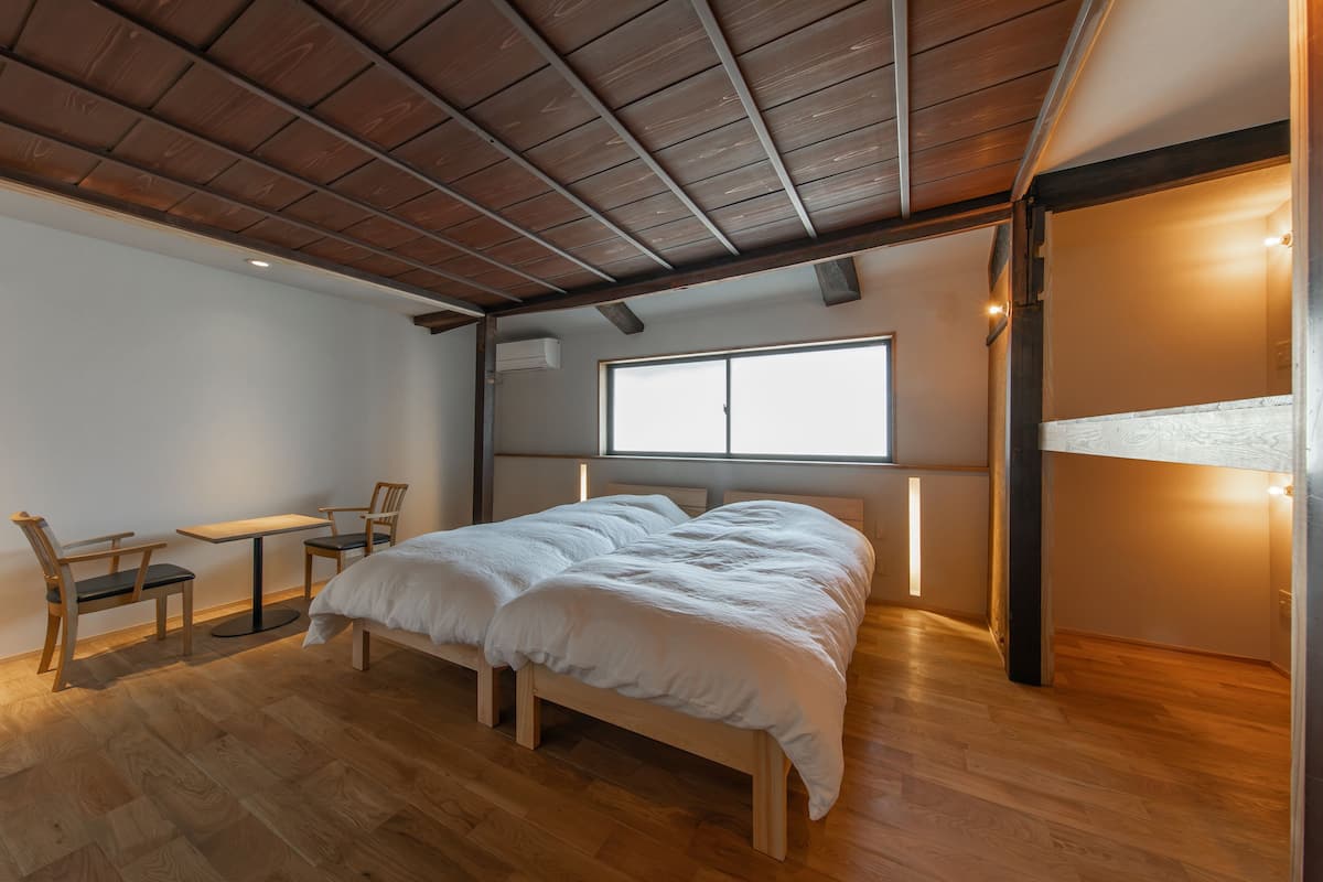 Ein Schlafzimmer mit Holzboden, einem Bett mit weißer Bettwäsche, einem kleinen Tisch mit Stühlen und einem Fenster, das natürliches Licht hereinlässt.