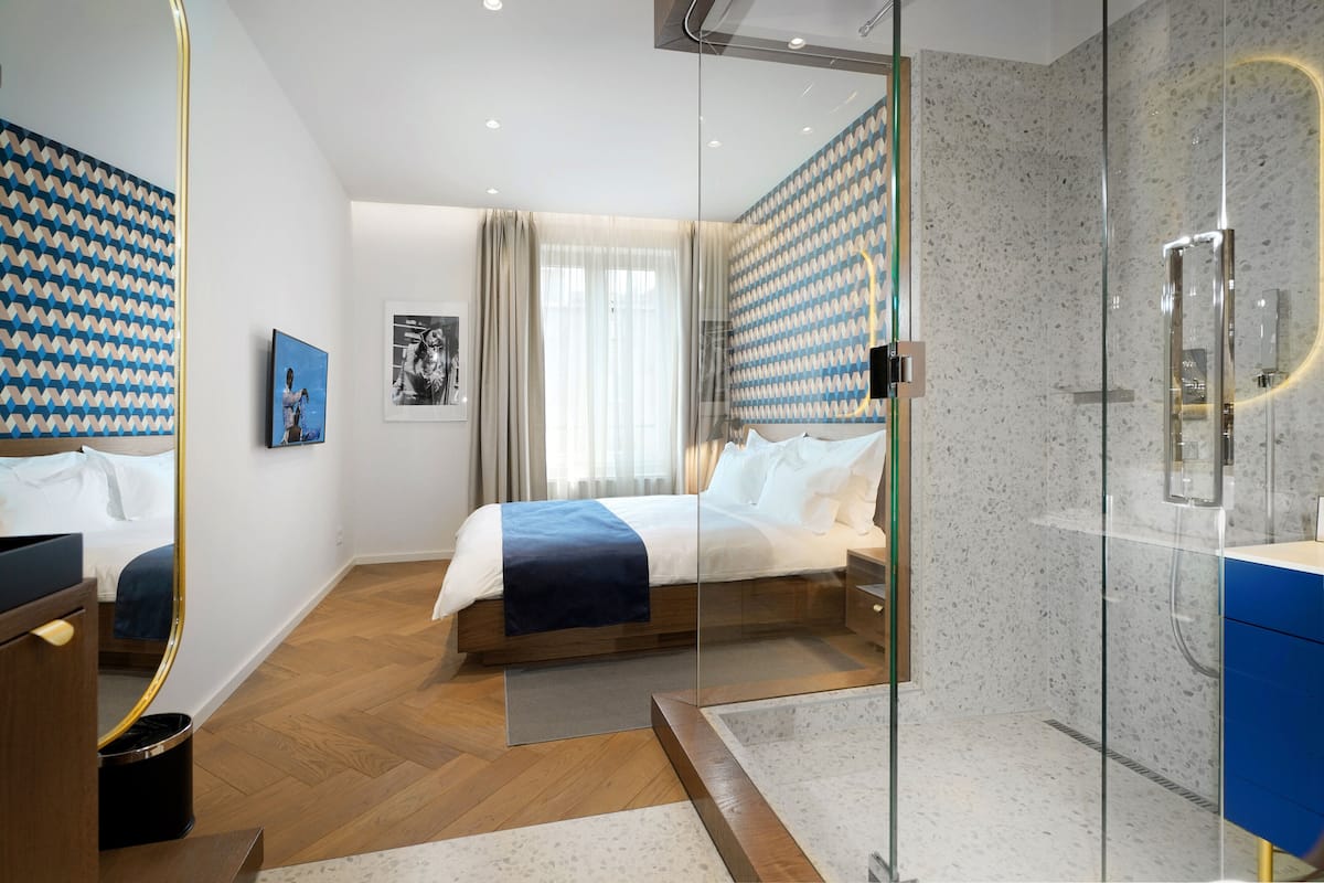 A modern hotel room with a large bed, a glass shower enclosure, and a wooden floor.