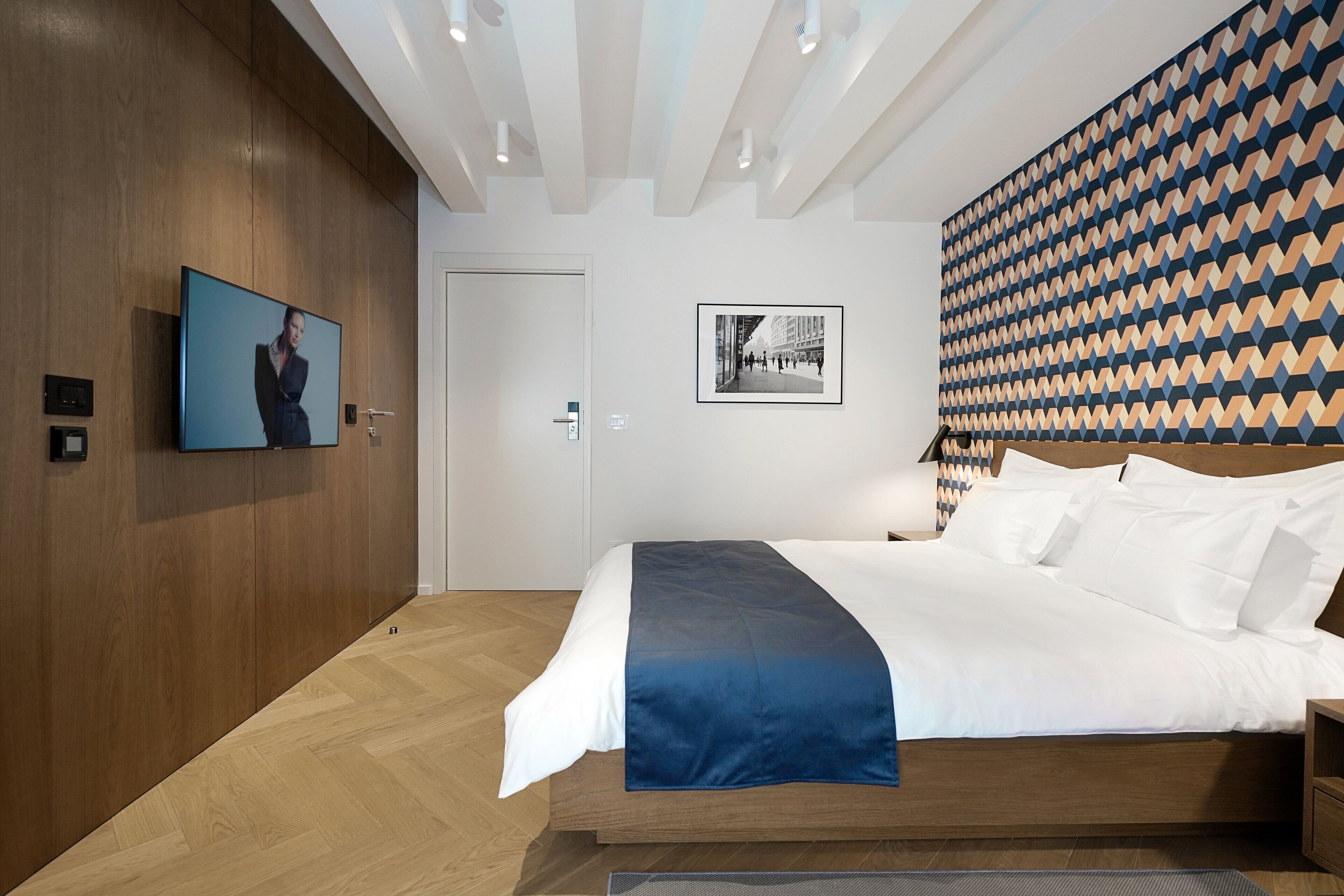 A modern hotel room with a wooden floor, a large bed with white and blue bedding, a flat-screen TV mounted on the wall, and a patterned wall design.