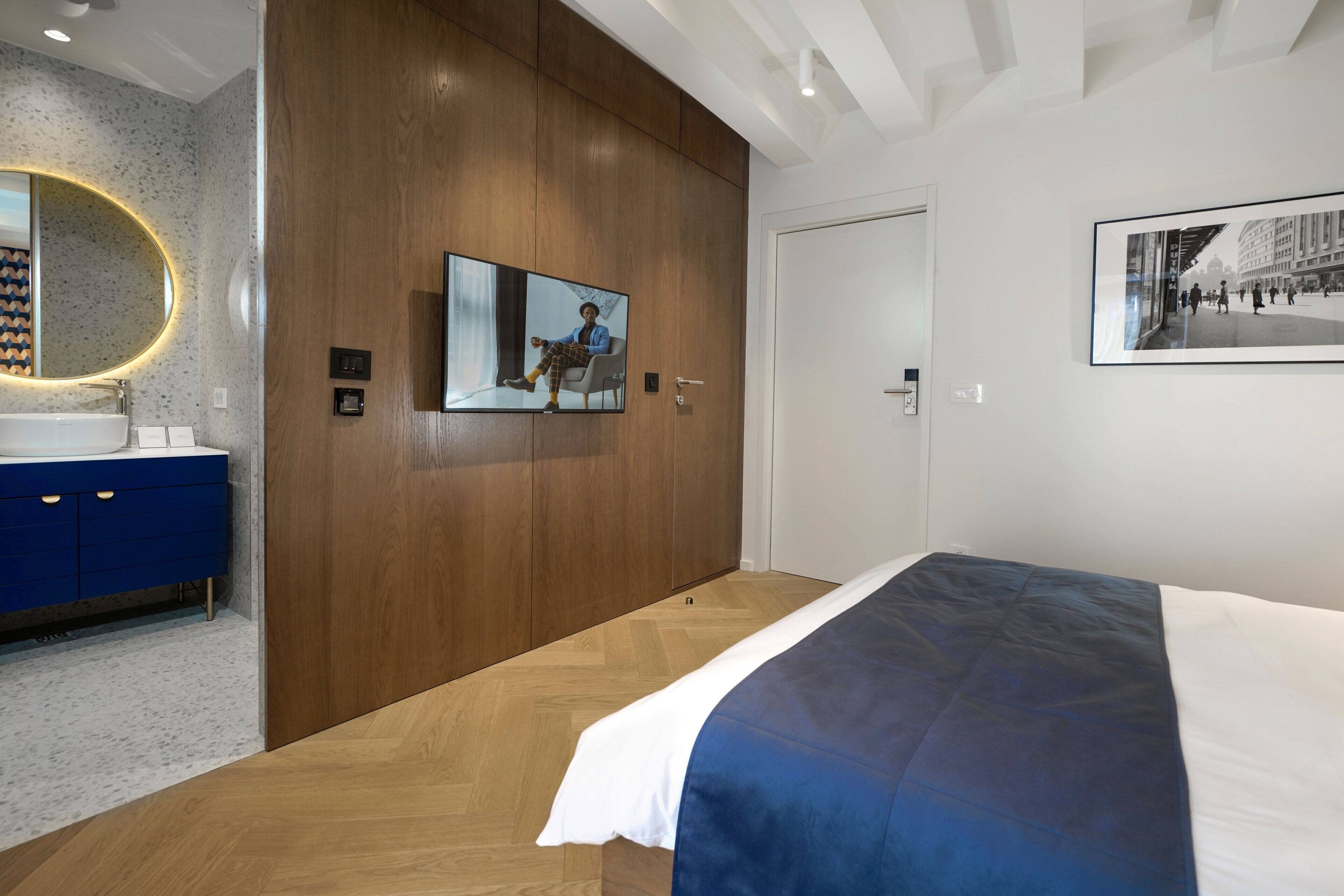 Un dormitorio moderno con un armario de madera, una cama grande con edredón azul y blanco, un espejo redondo con un halo de luz, y un cuadro enmarcado en la pared.