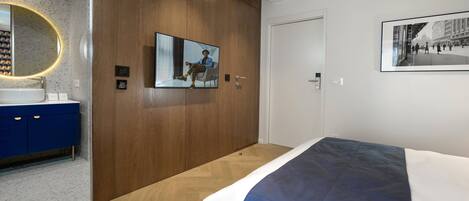 Un dormitorio moderno con un armario de madera, una cama grande con edredón azul y blanco, un espejo redondo con un halo de luz, y un cuadro enmarcado en la pared.