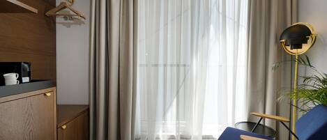 Ein modernes Hotelzimmer mit einem Holzschrank, einem blauen Sessel, einem runden Spiegel und leichten Vorhängen.