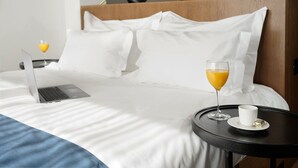 A neatly made bed with white linens, a laptop, a glass of orange juice, and a cup on a small table beside it. - Dominic Smart & Luxury Suites - Edition (Belgrade)