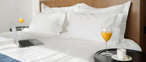 Una cama bien tendida con sábanas blancas, una computadora portátil, un vaso de jugo de naranja y una taza sobre una mesita de noche.