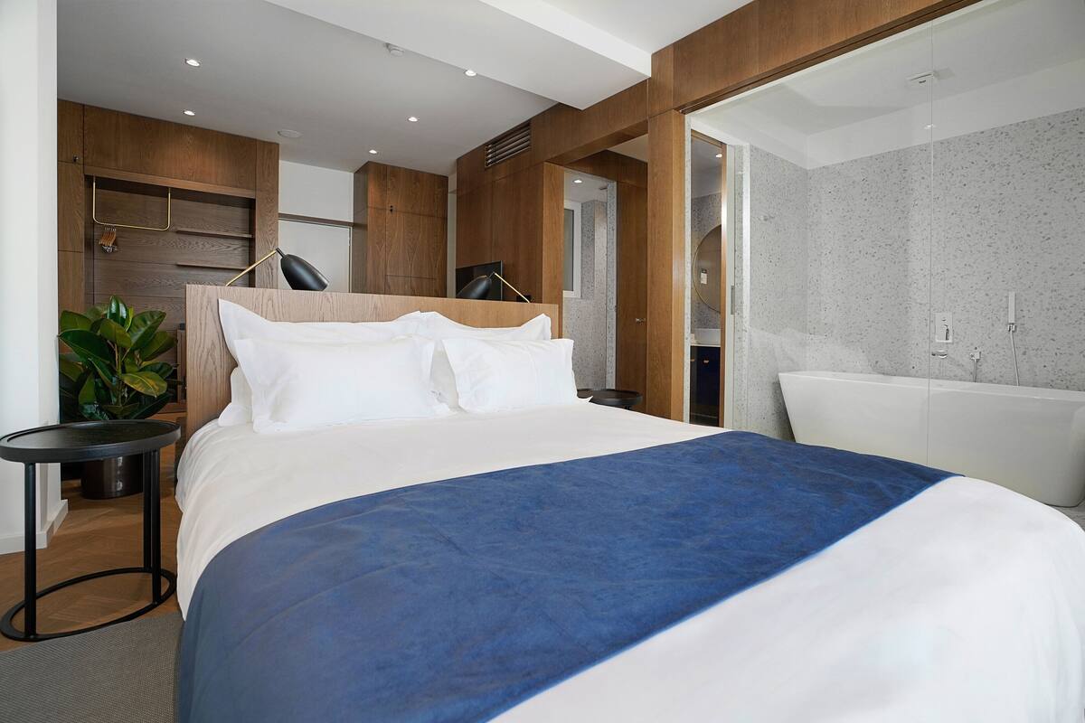 A modern hotel room with a large bed, a small round table, a potted plant, and a view into the bathroom.