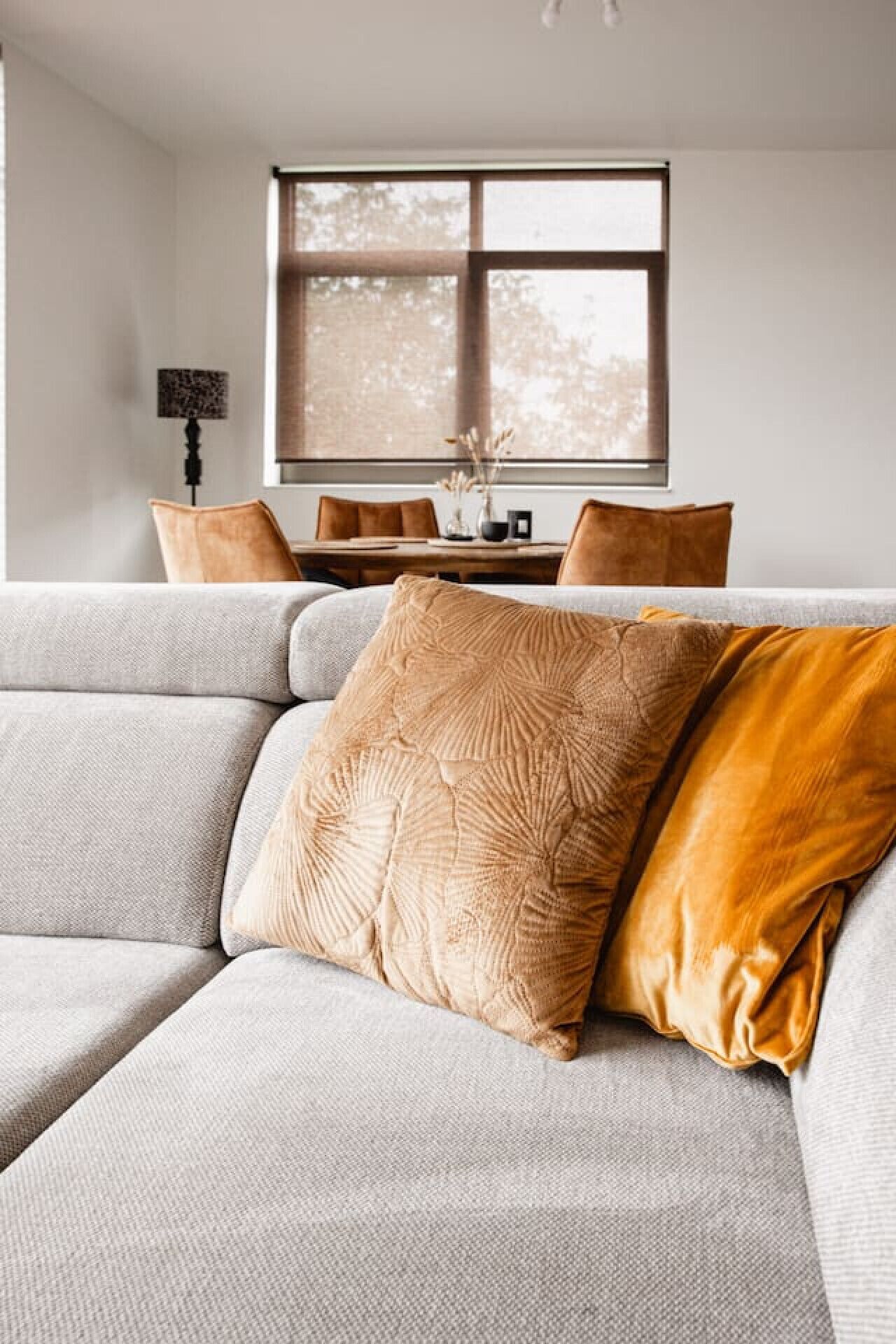 A modern living room with a grey sofa, a large window with blinds, and a dining area with a table and chairs.