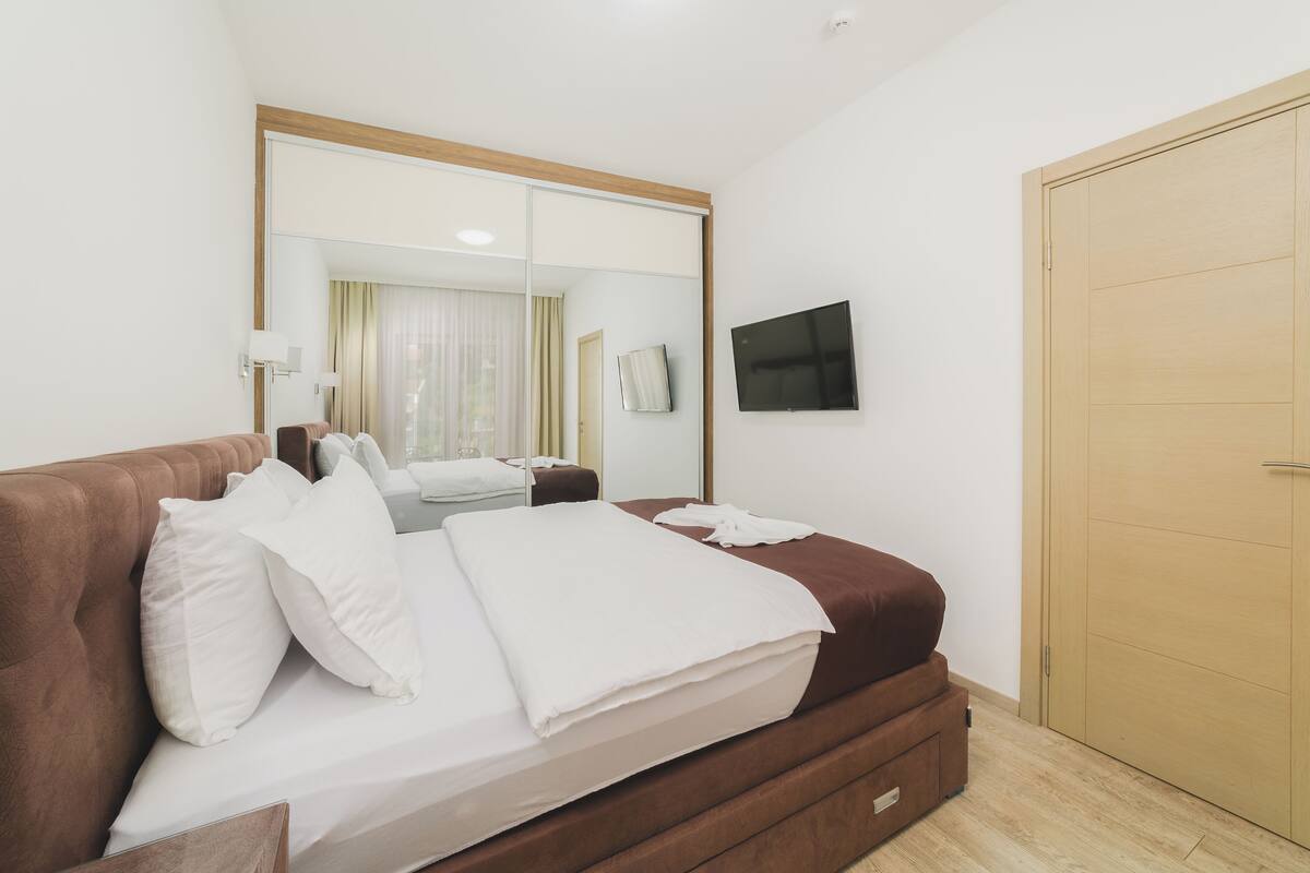 A modern hotel room with a large bed, a flat-screen TV, a wardrobe, and a mirror reflecting the room.