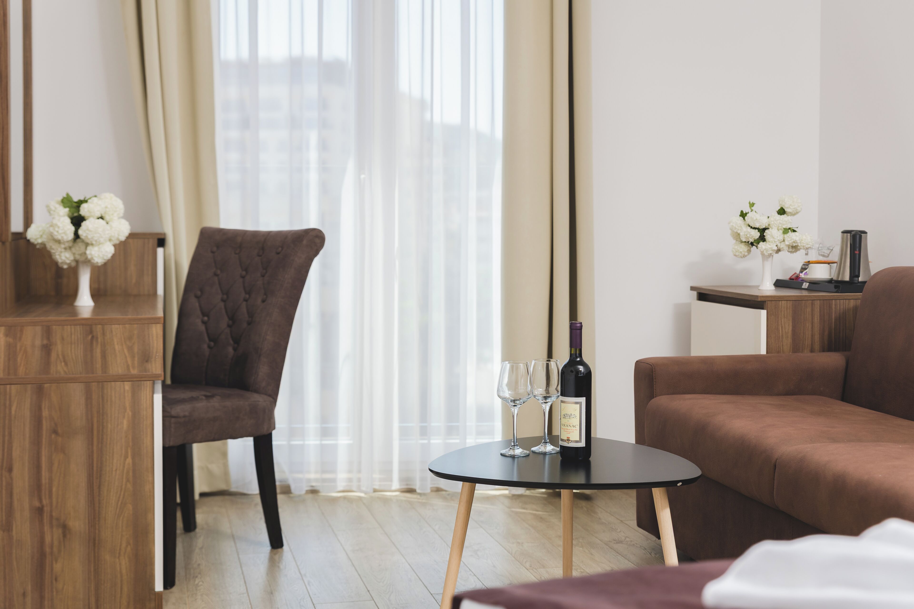 Habitación moderna con un sofá tapizado marrón, una mesa redonda negra con una botella de vino y dos copas, un sillón capitoné y un pequeño jarrón blanco con flores sobre una mesita de madera.