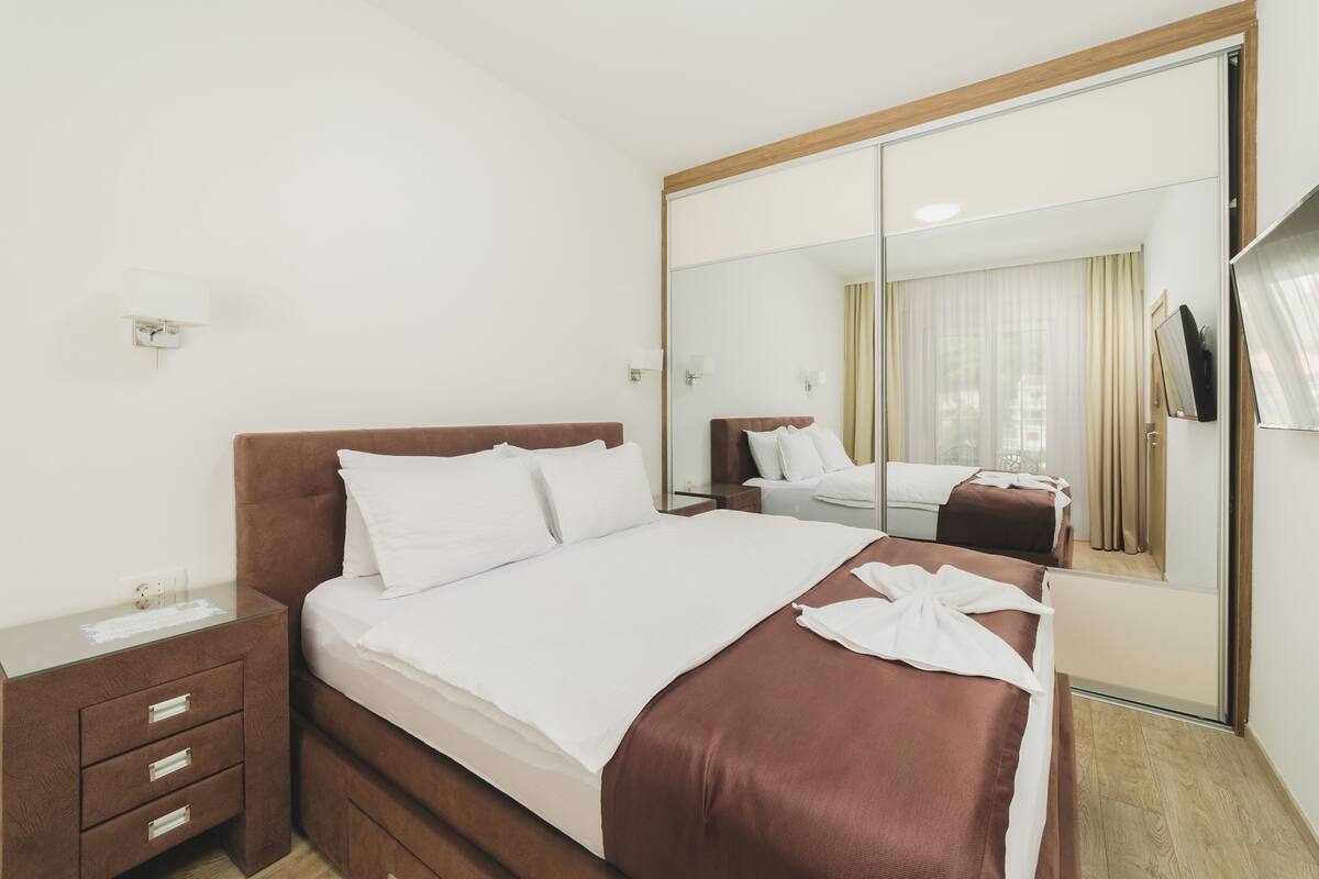 A hotel room with a large mirror, a bed with white and brown bedding, a wooden nightstand, and a small desk with a lamp.