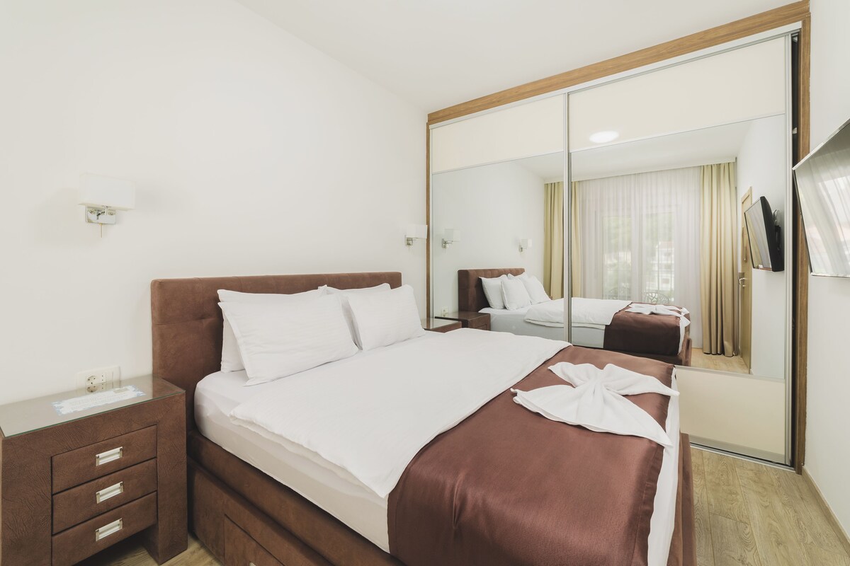A hotel room with a large mirror, a bed with white and brown bedding, a wooden nightstand, and a small desk with a lamp.