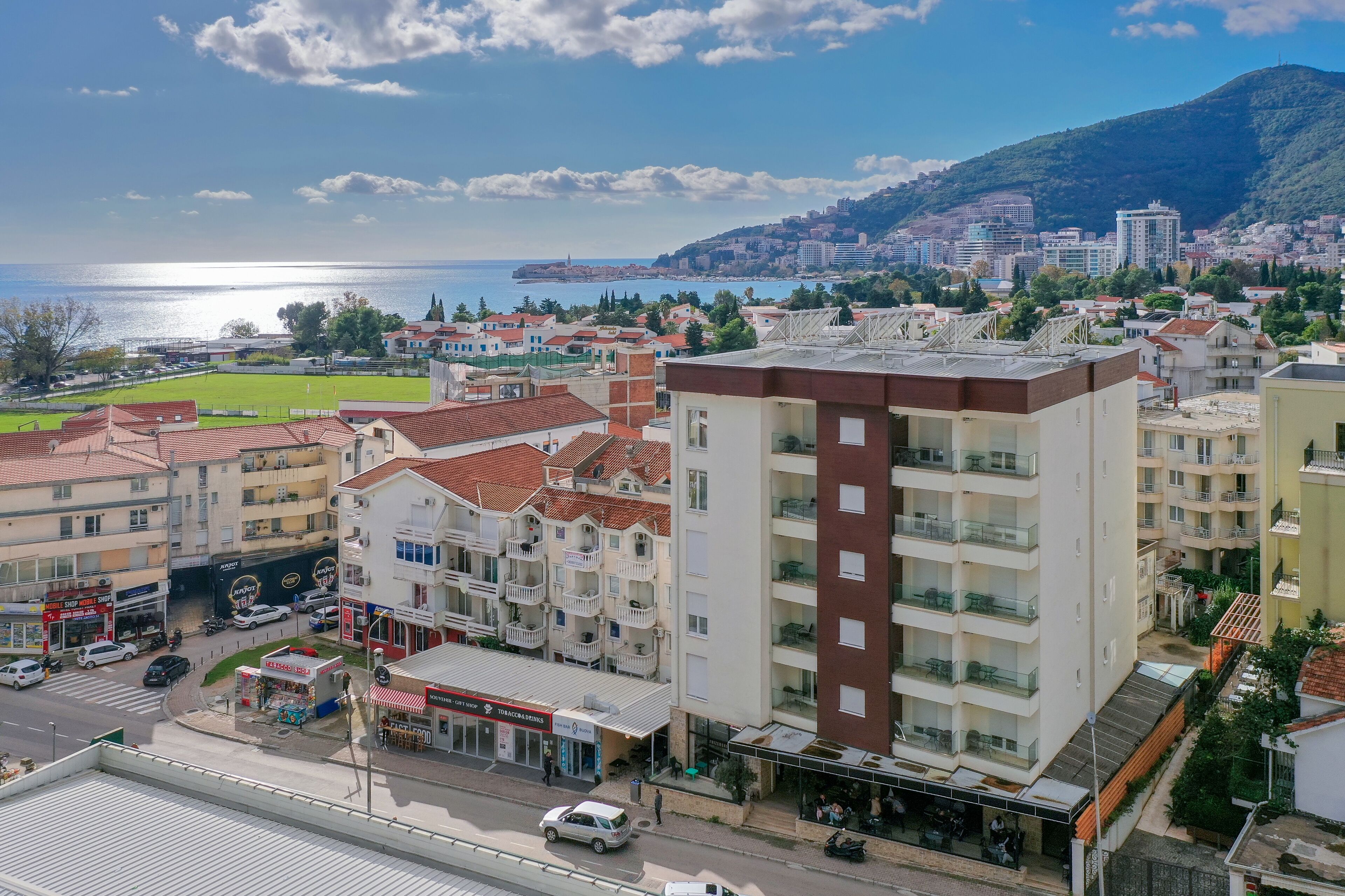 Un hotel de varios pisos con balcones, una calle visible al frente y vistas al mar y una colina al fondo.