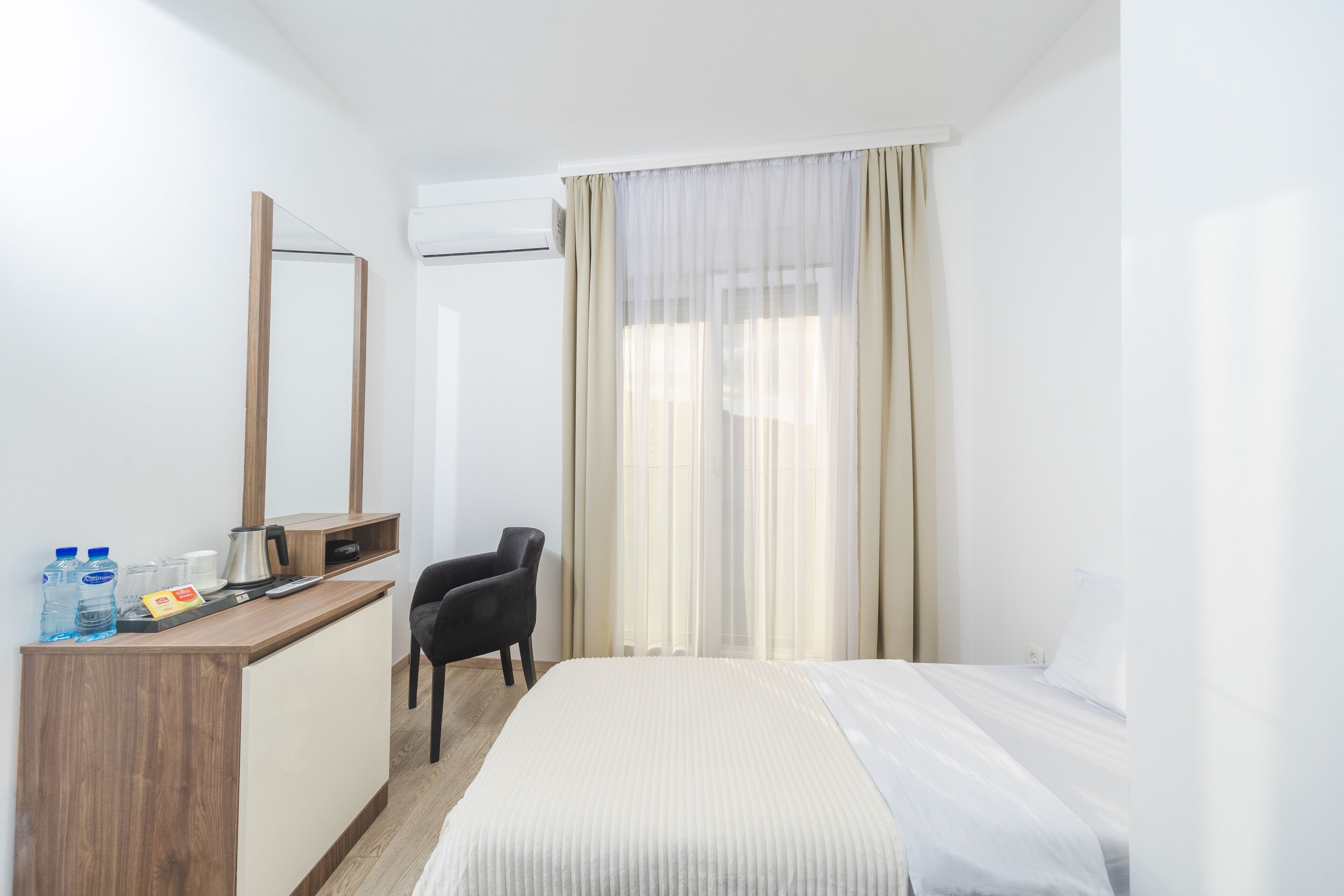 A hotel room with a bed, a desk with a mirror, a chair, and a window with sheer curtains.