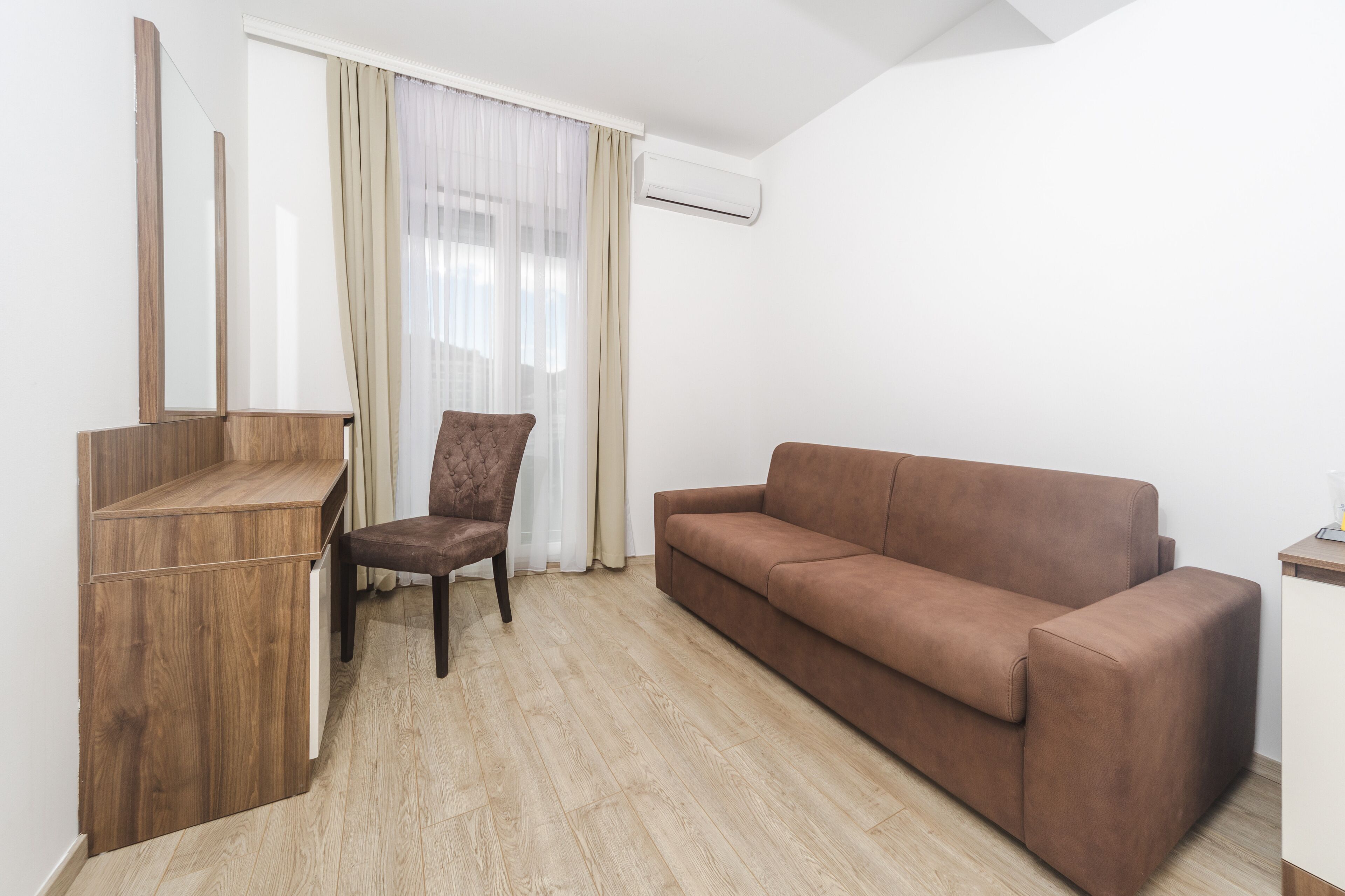 A room with a wooden desk, a brown sofa, a chair, and a window with sheer curtains.
