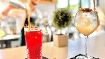 Two drinks on a table with a small bowl of nuts and a potted plant in the background.