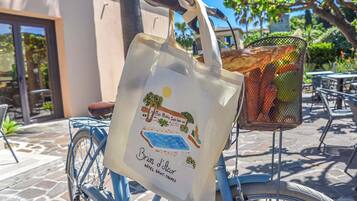 A bicycle with a basket containing a baguette and a wicker basket with fruits, parked on a cobblestone path with a building in the background.