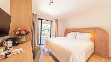 A hotel room with a bed, a desk with a computer, a vase of flowers, and a view of the outdoors.