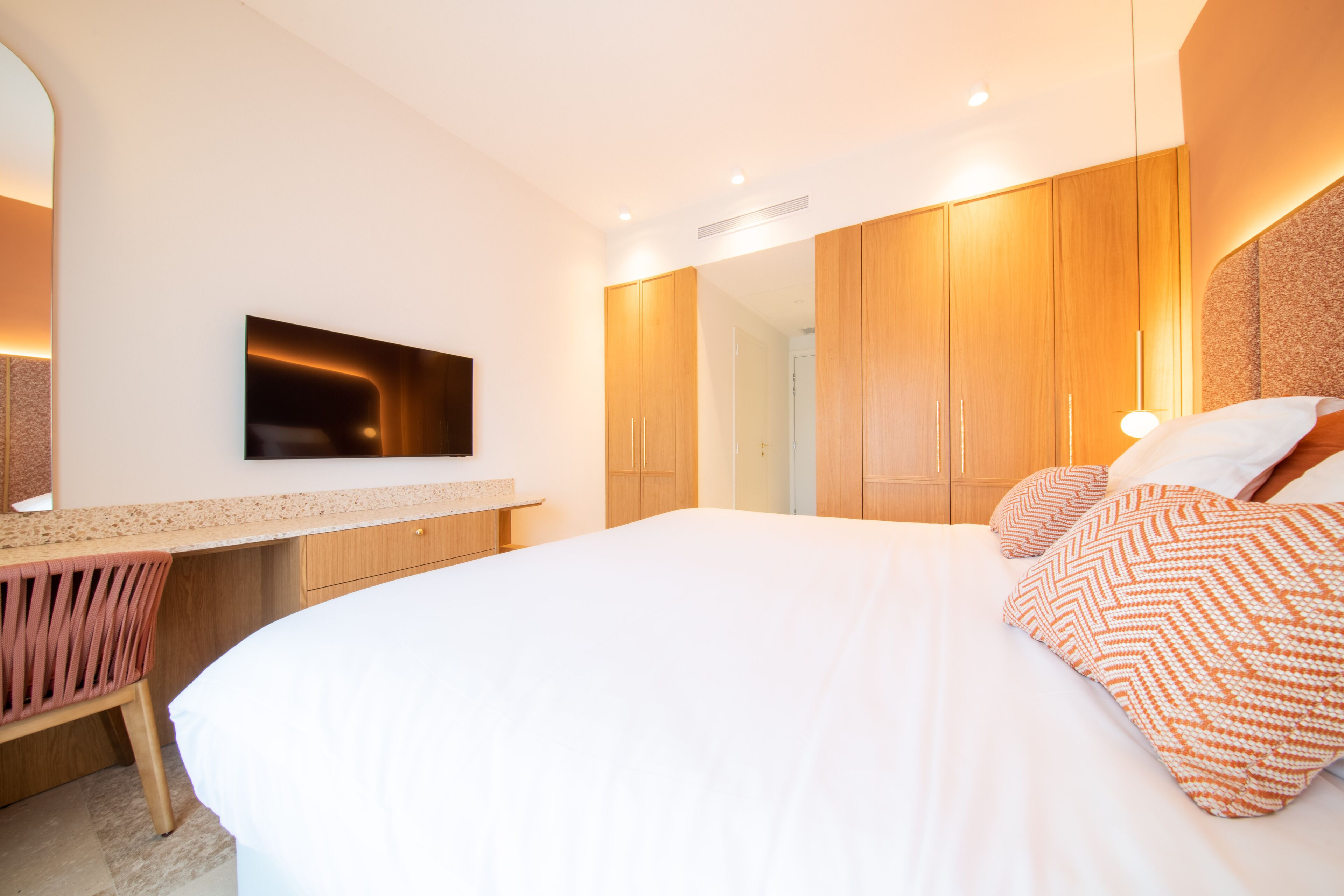 A modern bedroom with a large bed, a built-in TV, wooden wardrobe, and a small desk with a chair.