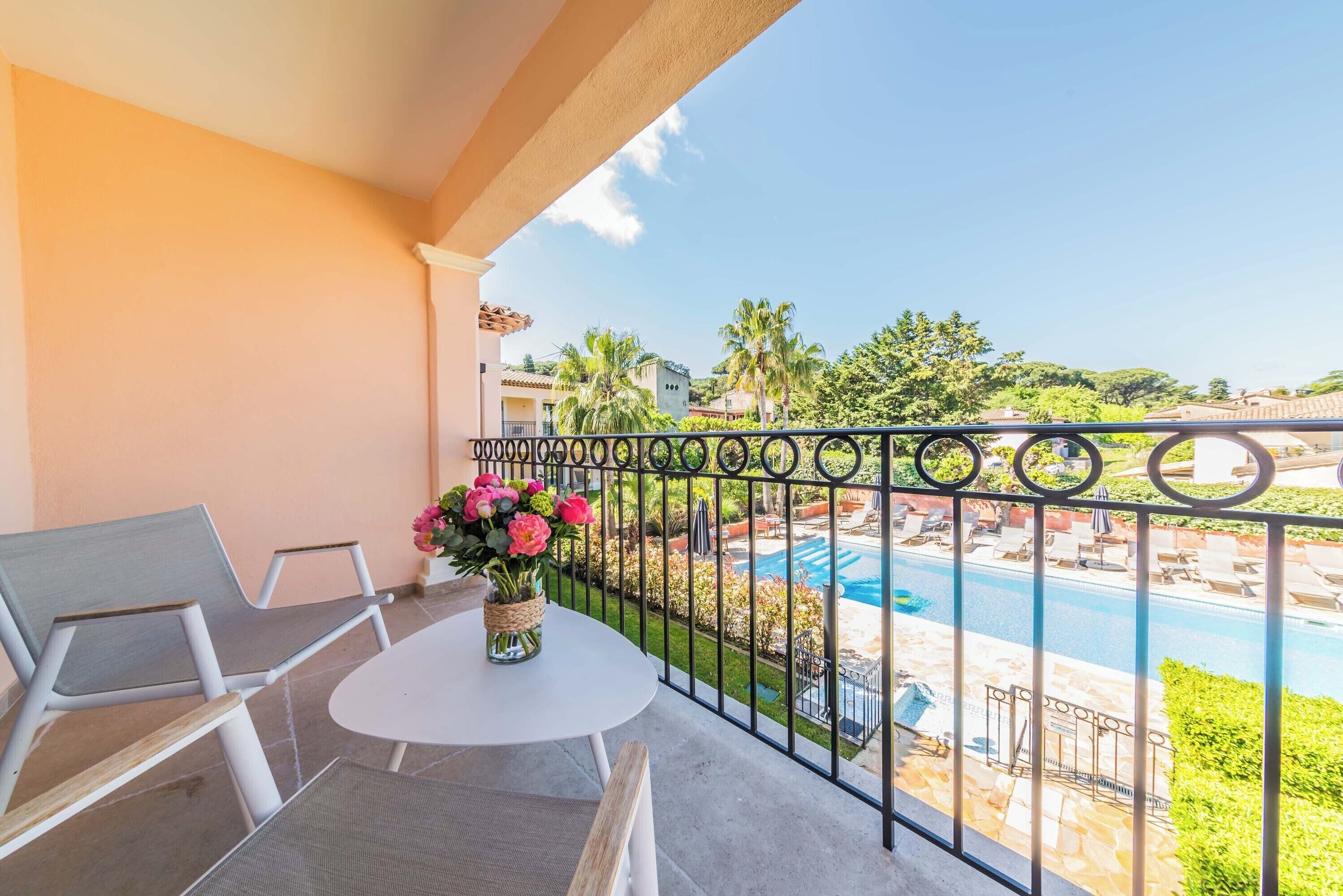 Un balcón con mesa y sillas blancas, un florero con flores y vista a una piscina y vegetación.