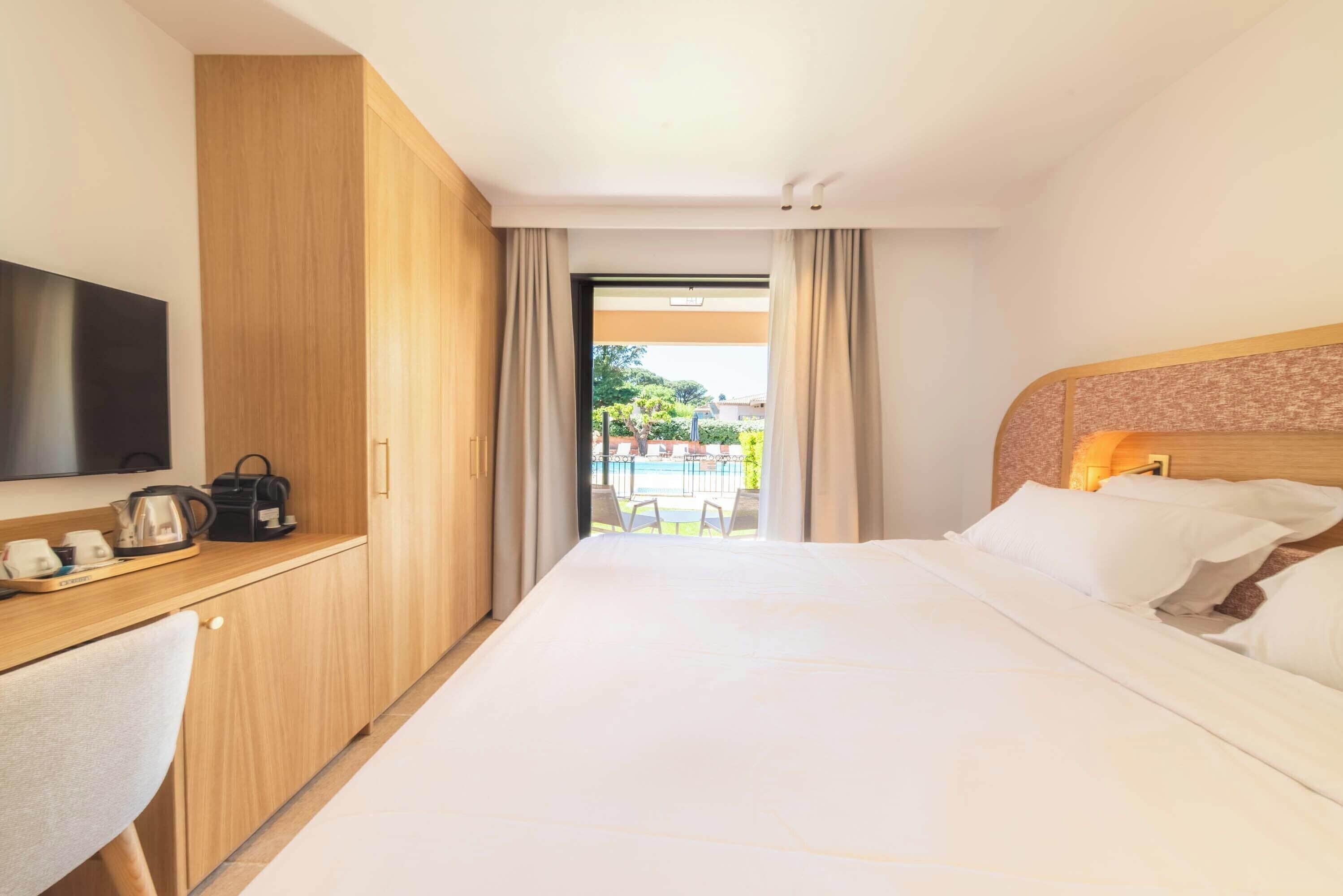 A hotel room with a bed, a desk with a television, a coffee maker, and a view of an outdoor pool area.