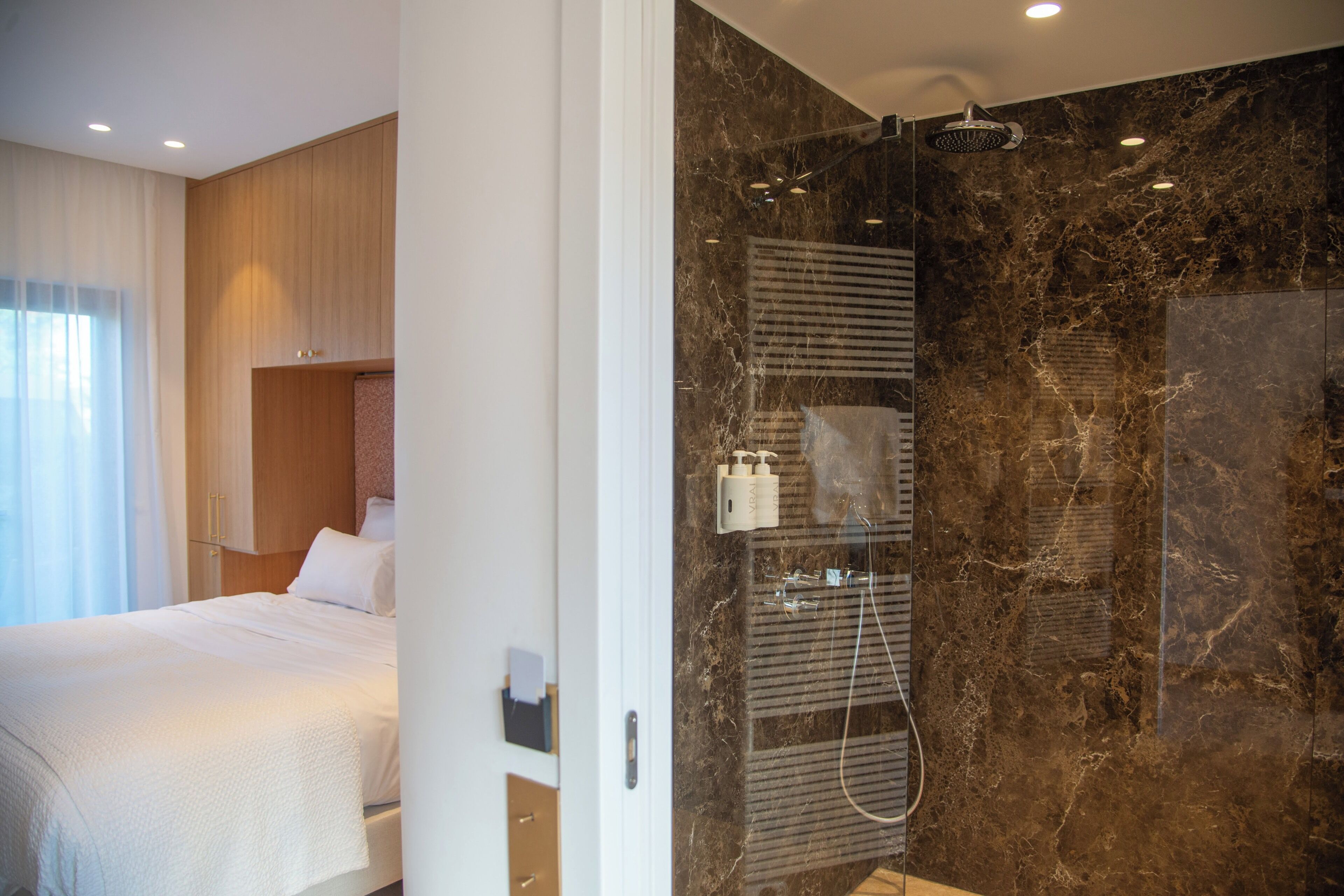 Habitación de hotel con cama, ducha con mampara de vidrio y pared de mármol.
