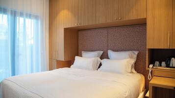 A hotel room with a bed, wooden headboard, white bedding, a bedside table with a phone, and a window with sheer curtains.
