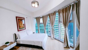 Un dormitorio con una cama grande, una lámpara colgante, un cuadro enmarcado y vistas de edificios altos a través de las ventanas.