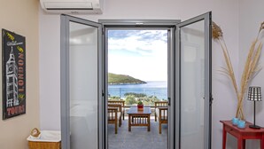 Una habitación con una puerta corrediza de vidrio que da a un balcón con vista al mar y una colina. En el balcón hay sillas de madera y una mesa, además de una mesa roja con una lámpara y objetos decorativos en el suelo.