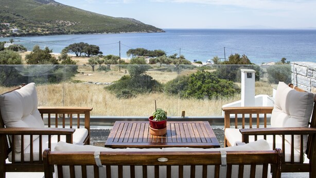 Un balcón con mobiliario de madera y una planta en maceta, con vistas a un paisaje costero con colinas y el mar.