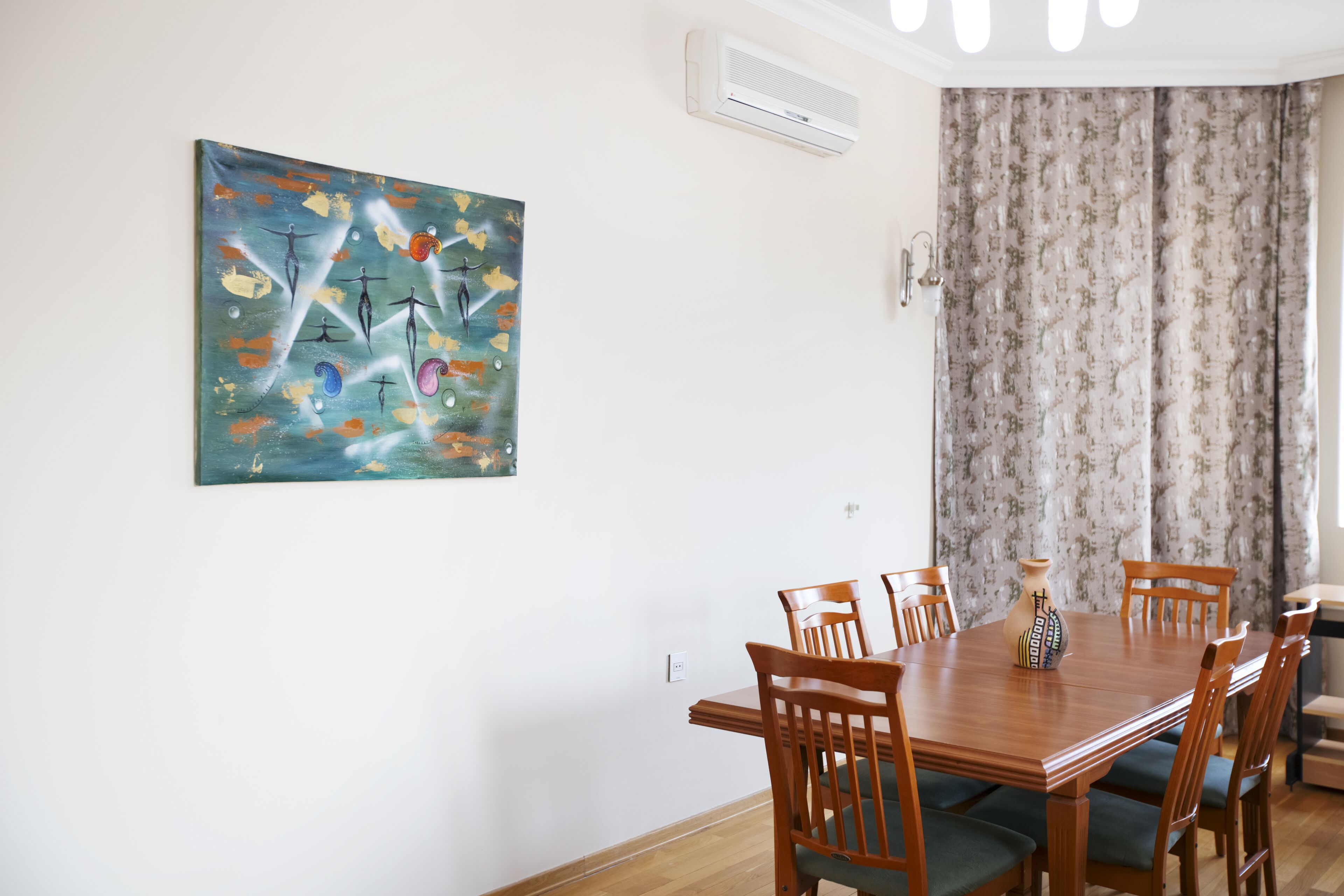 A dining area with a wooden table and chairs, a painting on the wall, and a window with curtains.