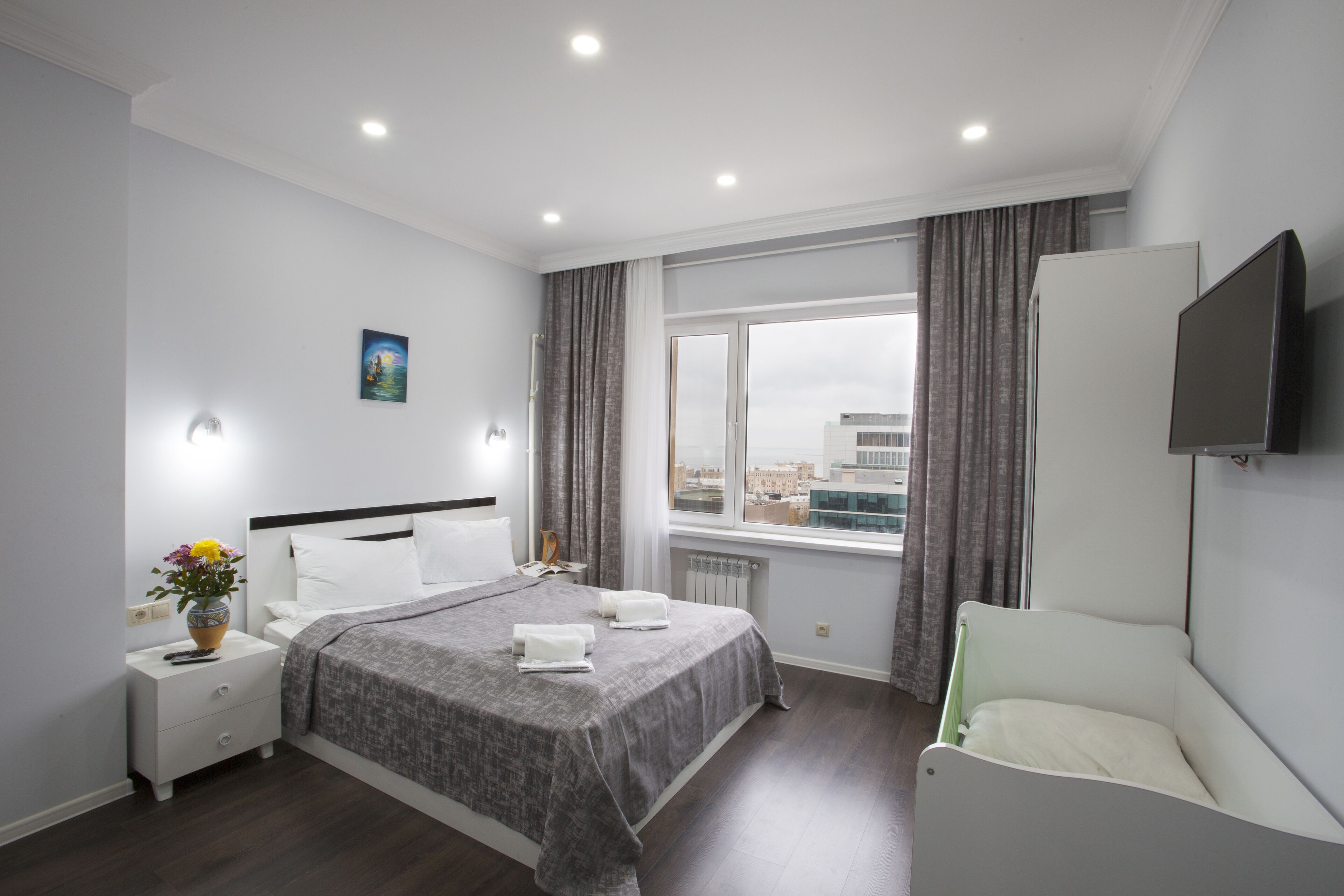A modern hotel room with a large bed, bedside tables, a TV, and a view of the city through the window.