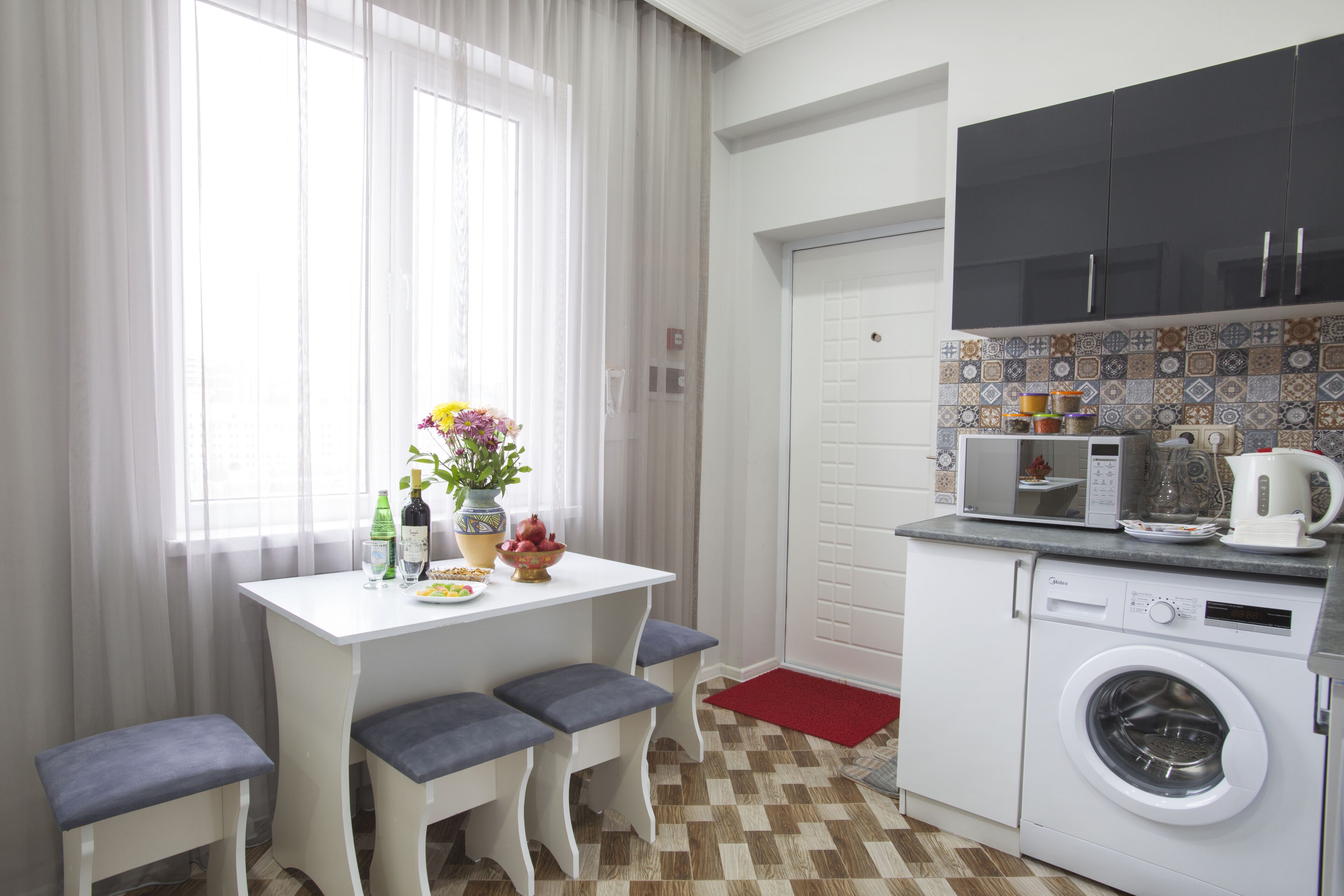A compact kitchen with a washing machine, a dining table with benches, and a window with sheer curtains.