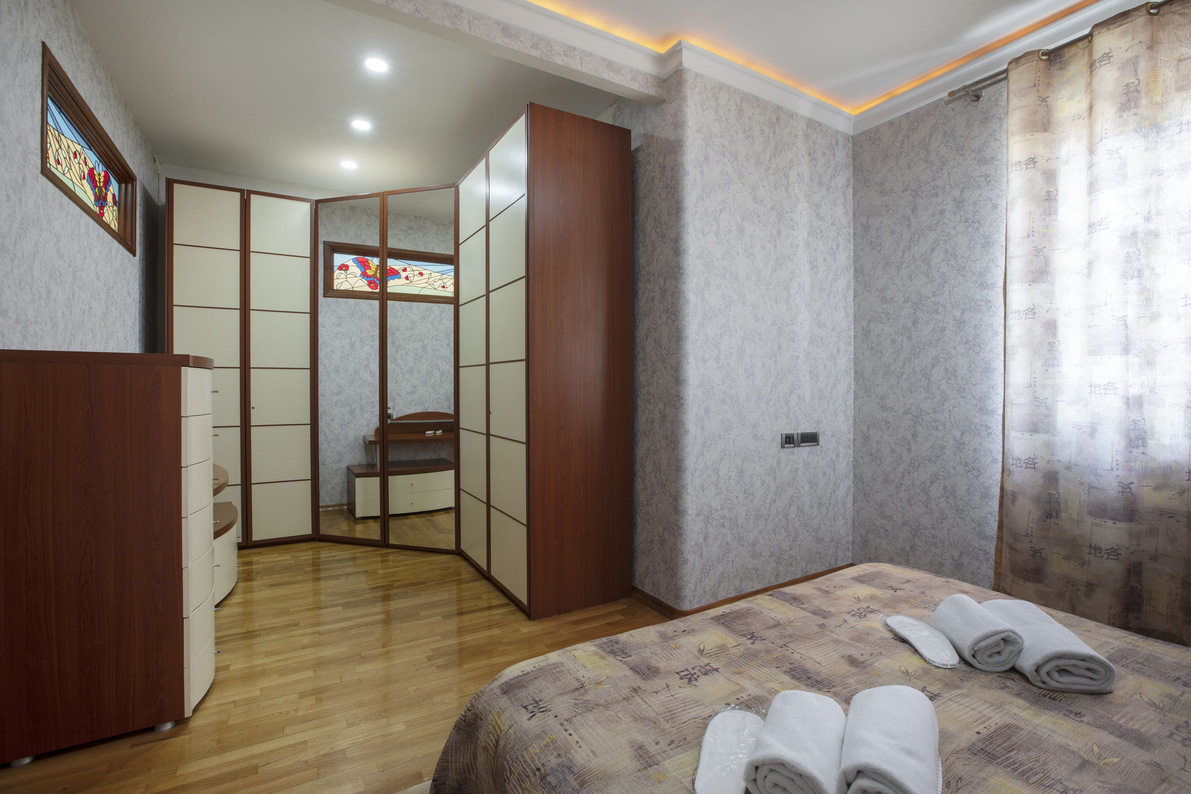 A hotel room with a bed, a wooden wardrobe, a small seating area, and a view of the outside through a window.