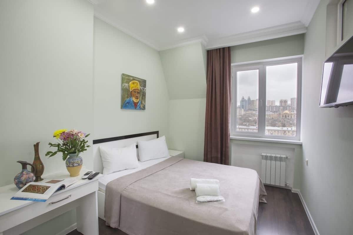 Une chambre d’hôtel avec un lit, une table de chevet, un vase avec des fleurs, un tableau au mur, une fenêtre donnant sur la ville et un radiateur.