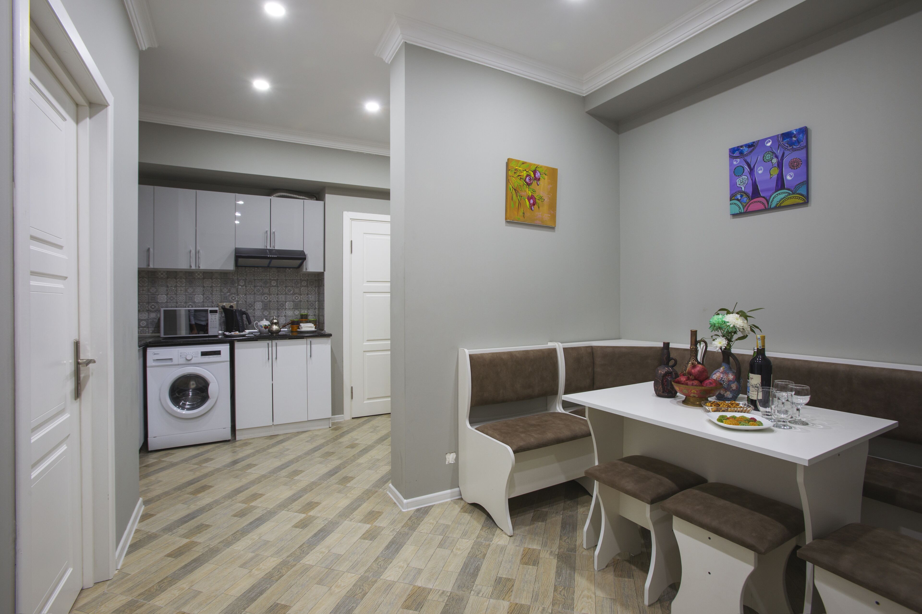 Une cuisine compacte équipée d’un lave-linge, un coin repas avec un banc et une table, et deux tableaux accrochés au mur.