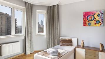 A hotel room with a large window, a bed with a patterned bedspread, a bedside table with a vase, and a painting on the wall.