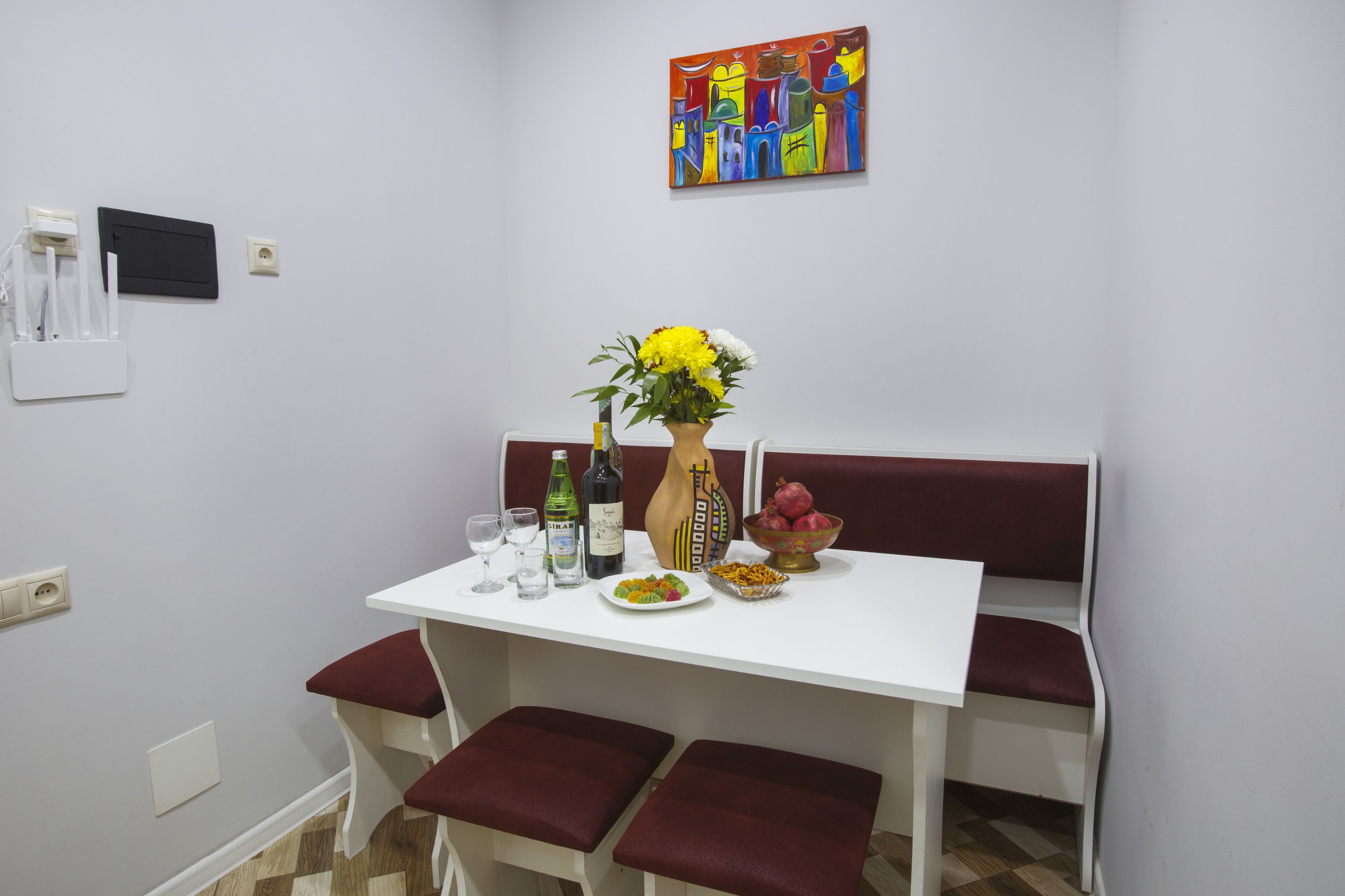 Un coin repas avec une table blanche, un banc rembourré de coussins rouges et un tableau coloré accroché au mur.