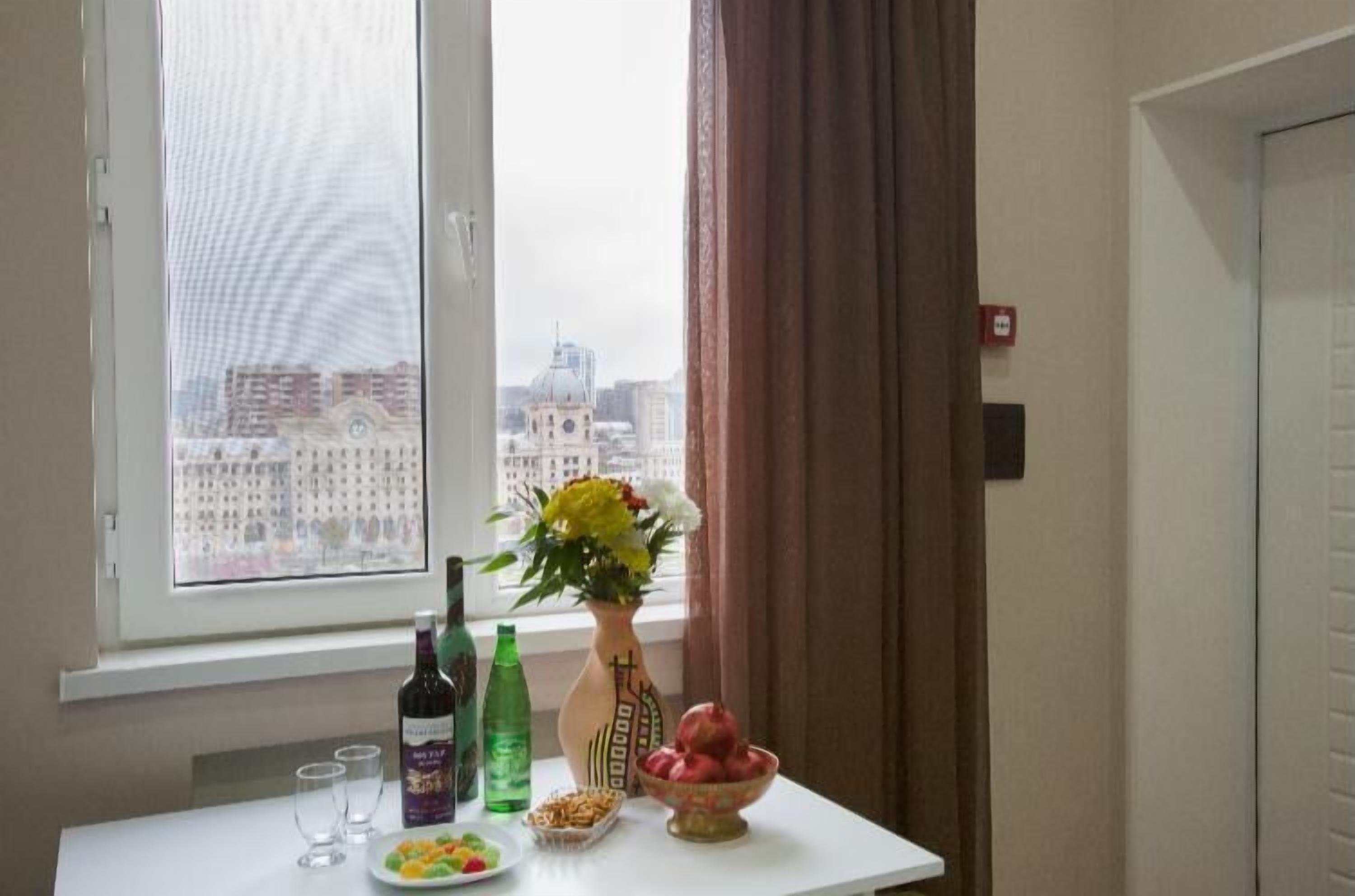 A table with a vase of flowers, a bottle of wine, and a bowl of fruit in a room with a window overlooking a cityscape.