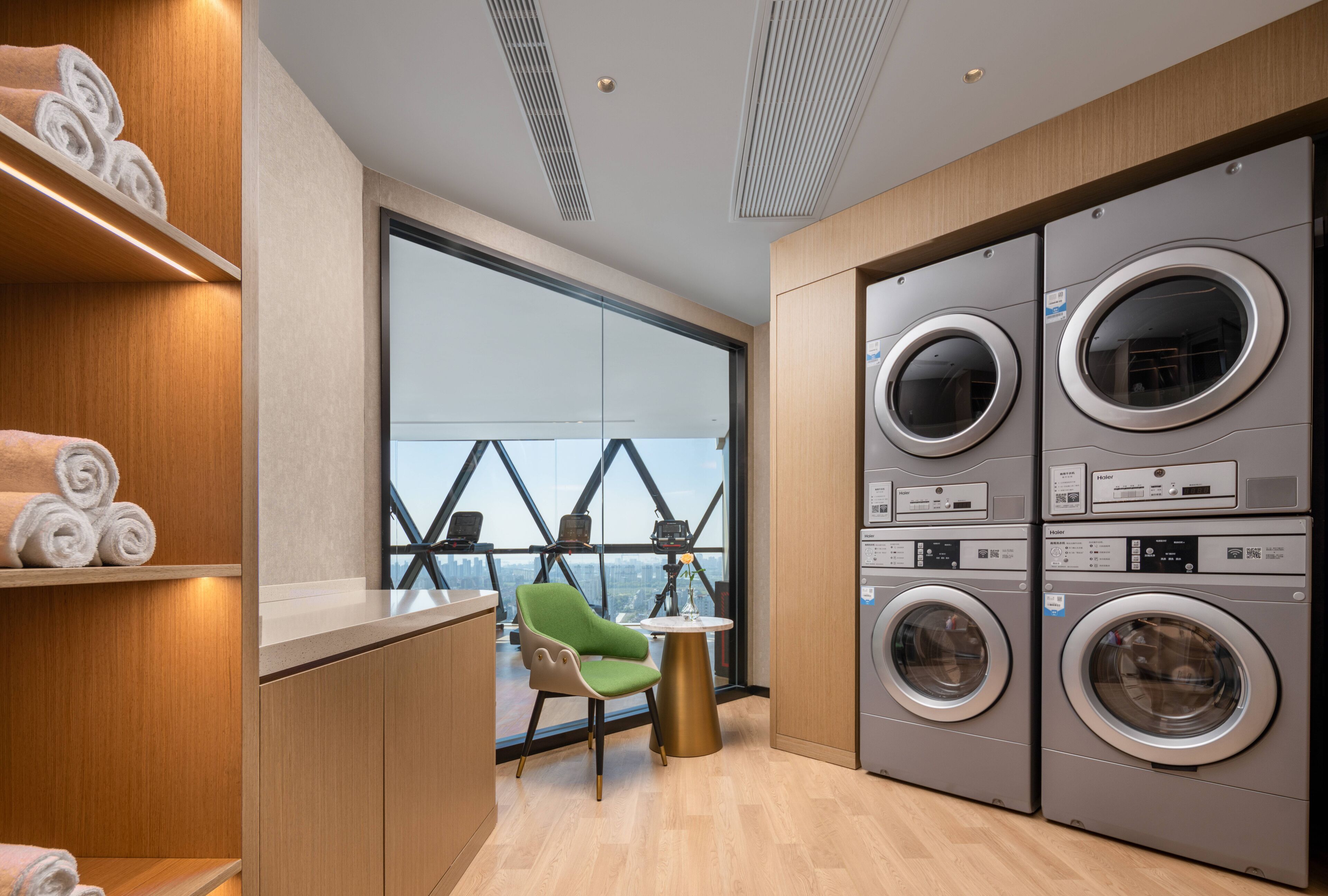 A modern laundry room with built-in washers and dryers, a green chair, and a view through large windows.